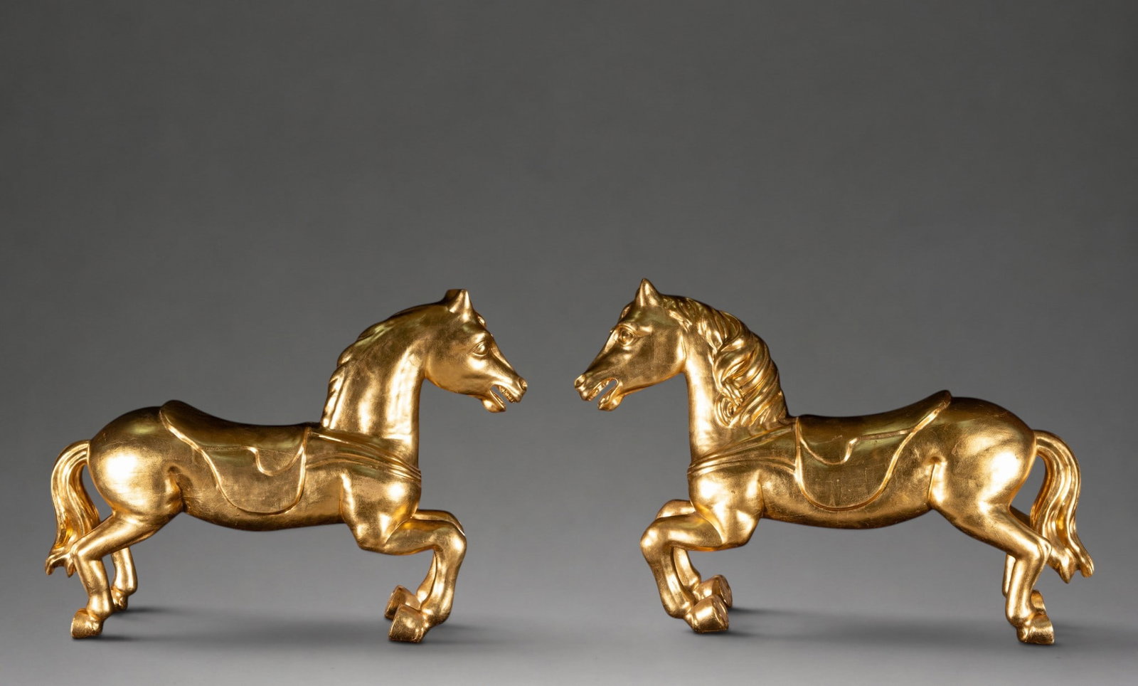 Pair of golden carousel horses – European School – first half of the 20th century: Pair of decorative sculptures made of carved, stuccoed, and gilded wood, modeled as facing carousel horses. They feature stylized anatomy, flowing manes, and saddle blankets or tack carved onto their 