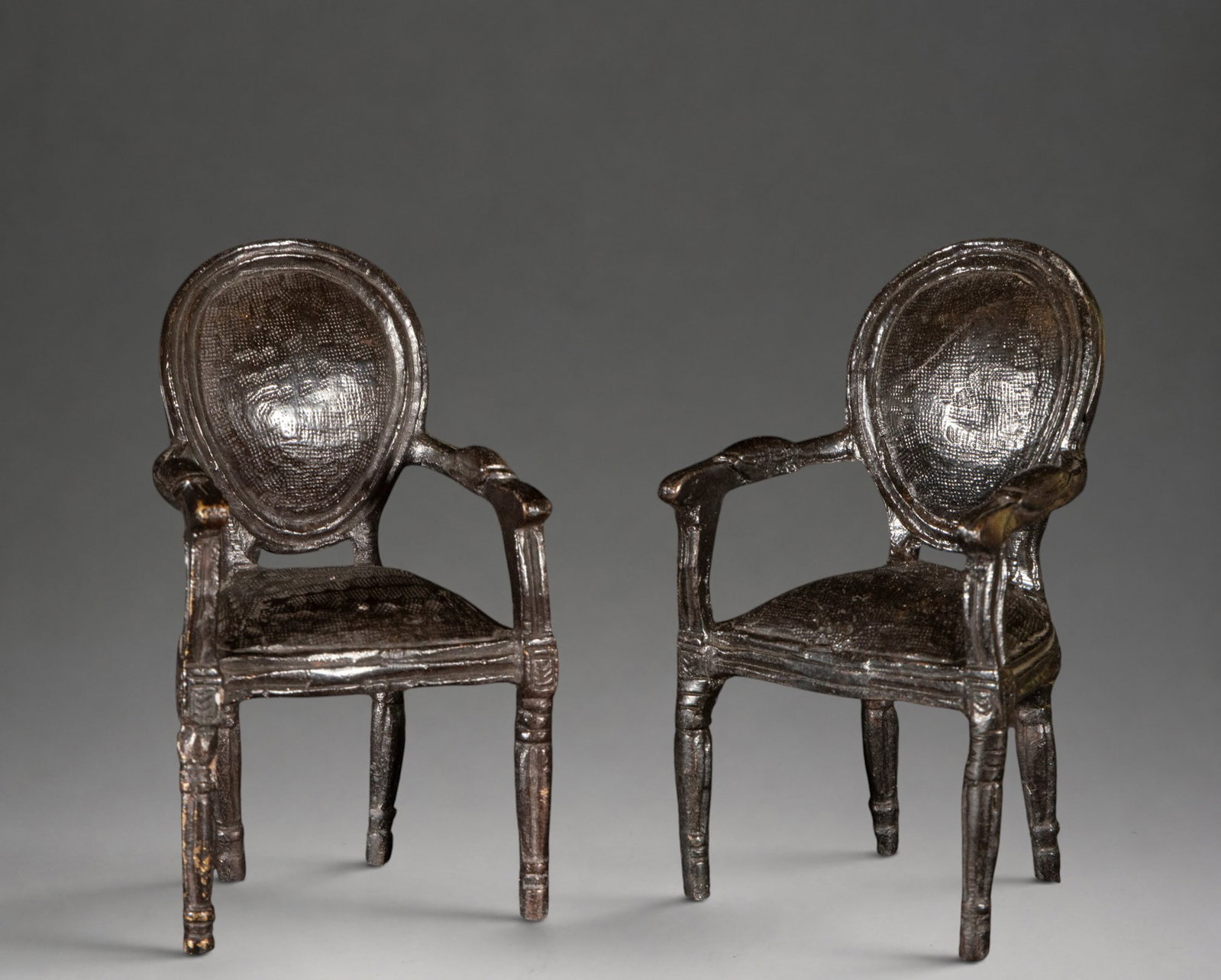 Pair of miniature Louis XVI style armchairs – 20th century: Patinated, cast, and chased bronze. Pair of miniature armchairs with oval backs, curved arms, and turned legs, inspired by French Louis XVI style models. The seat and back have textured surfaces that 