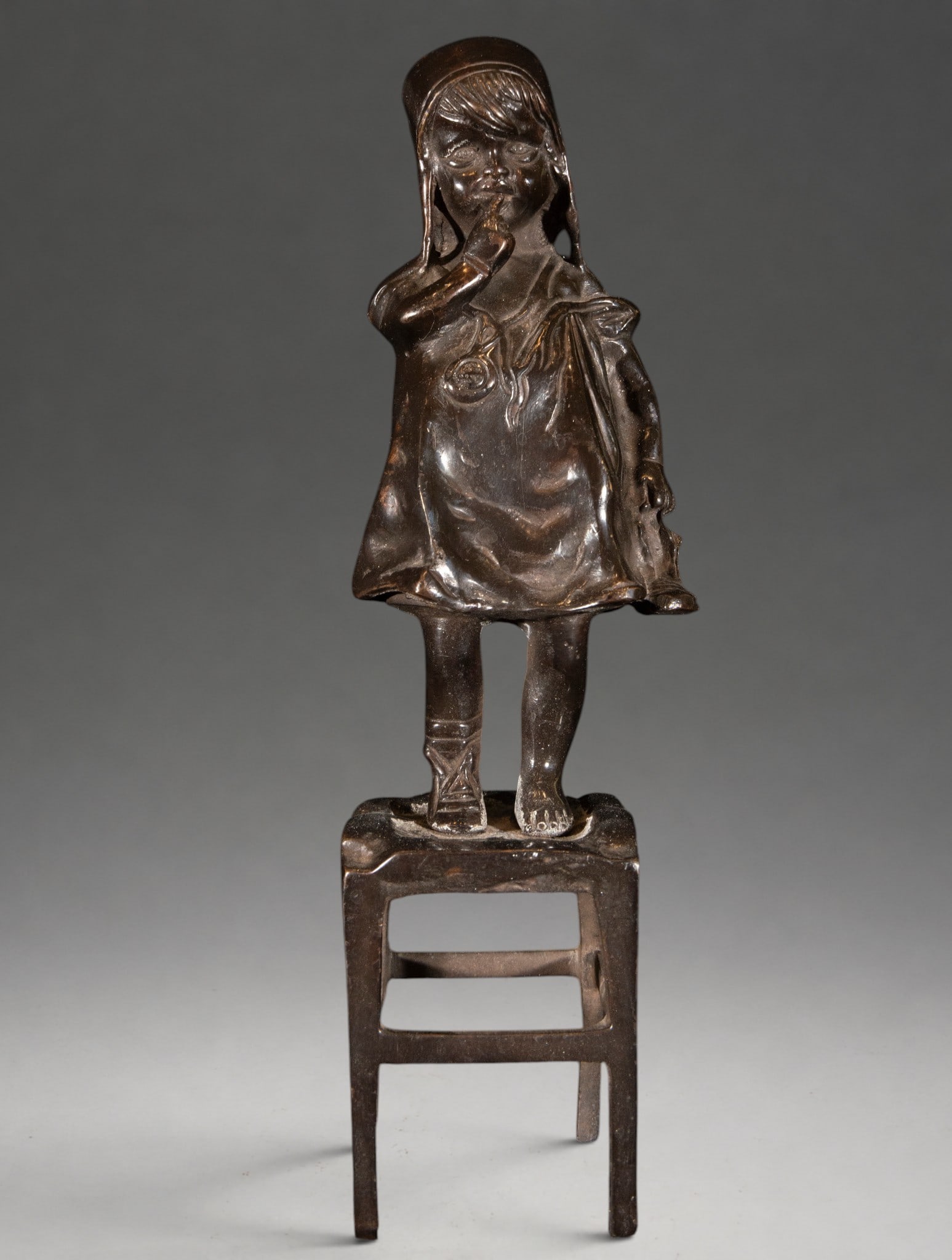 Girl on a chair – 20th century: Patinated bronze, cast. Child figure depicted standing on a chair, wearing folk-inspired clothing and a headdress that partially covers the hair. One hand is raised to the face while the other holds a