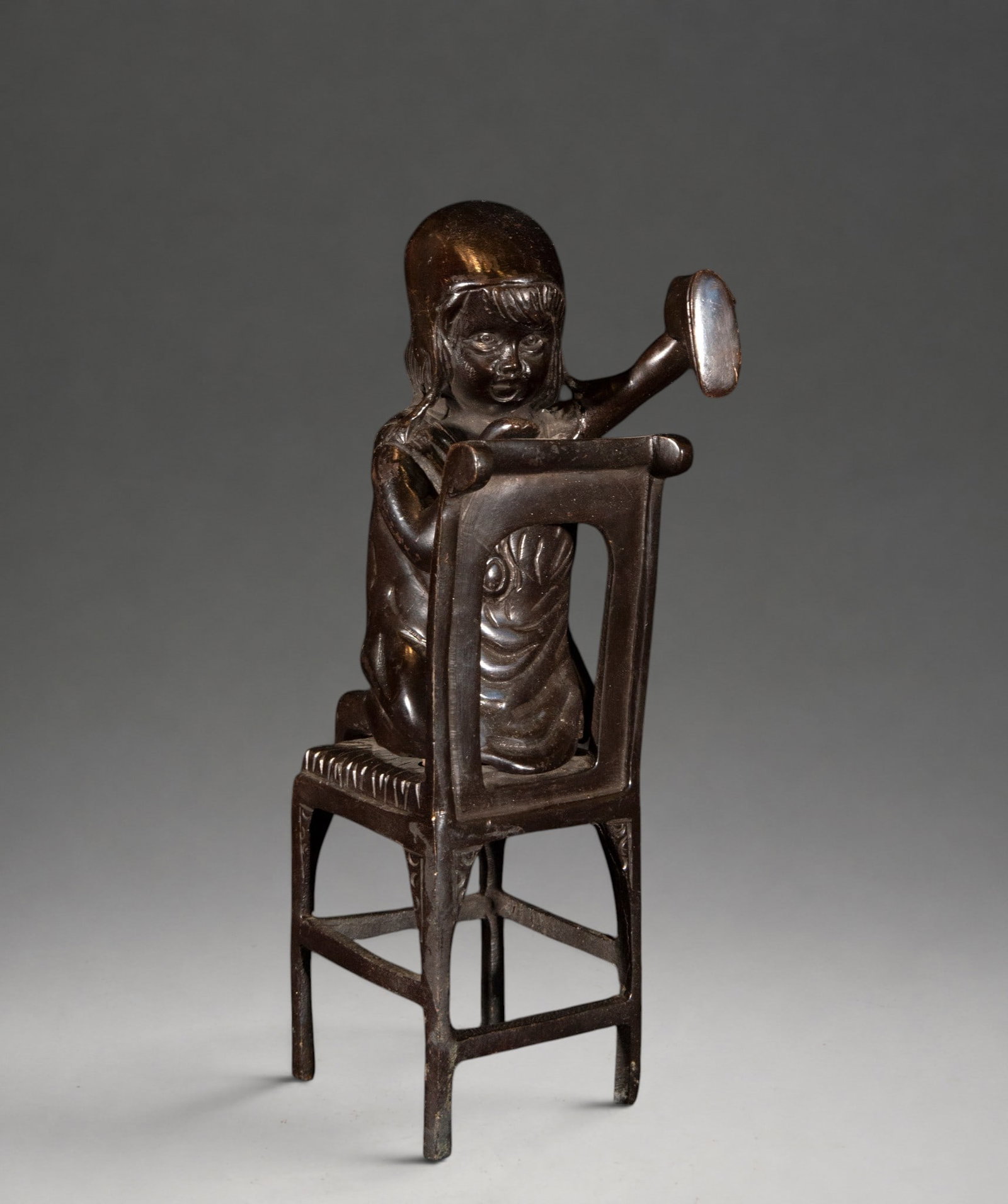 Girl sitting with mirror – 20th century: Patinated bronze, cast. Child figure seated on a chair, holding a small circular mirror with its hand raised. The figure features stylized modeling of the face, hair, and folds of the clothing, integr