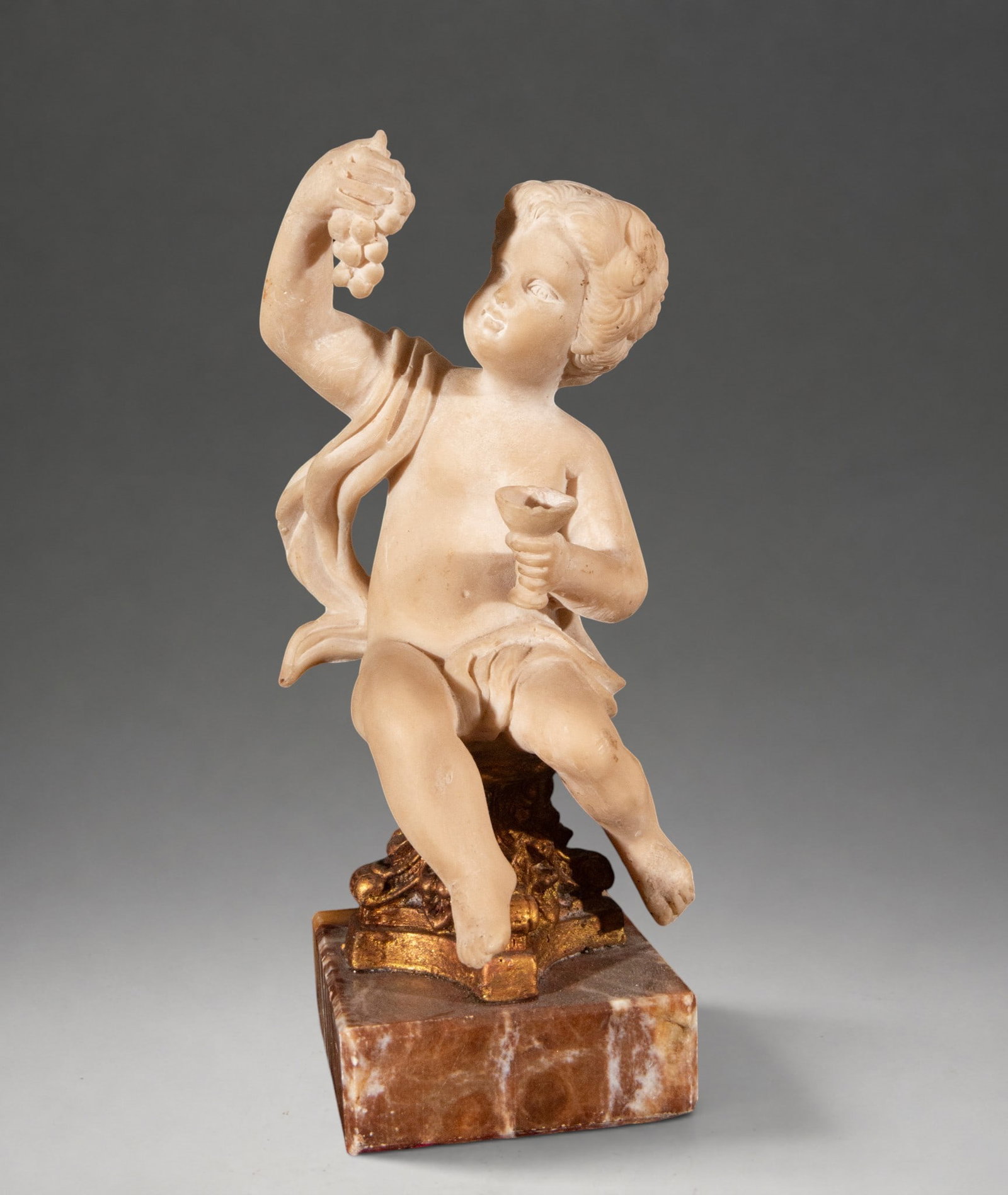 Bacchus as a child – European School – 19th century: Alabaster sculpture depicting Bacchus as a child, seated on an ornamental support and holding a raised bunch of grapes in one hand and a cup in the other, attributes alluding to the god of wine. The f