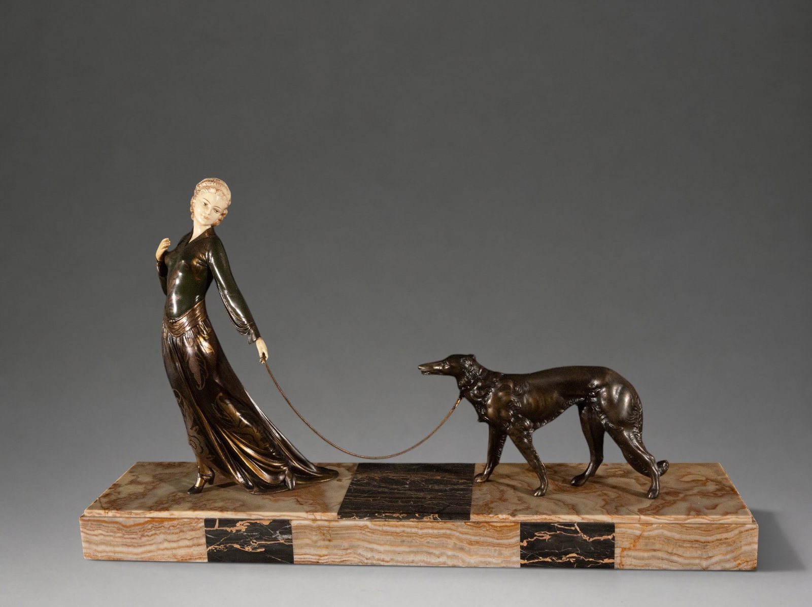 Lady with Greyhound – Chryselephantine Art Deco JR Colinet – European School – circa 1930: Art Deco sculptural group depicting an elegant female figure walking a greyhound. The figure combines patinated metal with light, ivory-like elements in the head and hands, and rests on a wide rectang