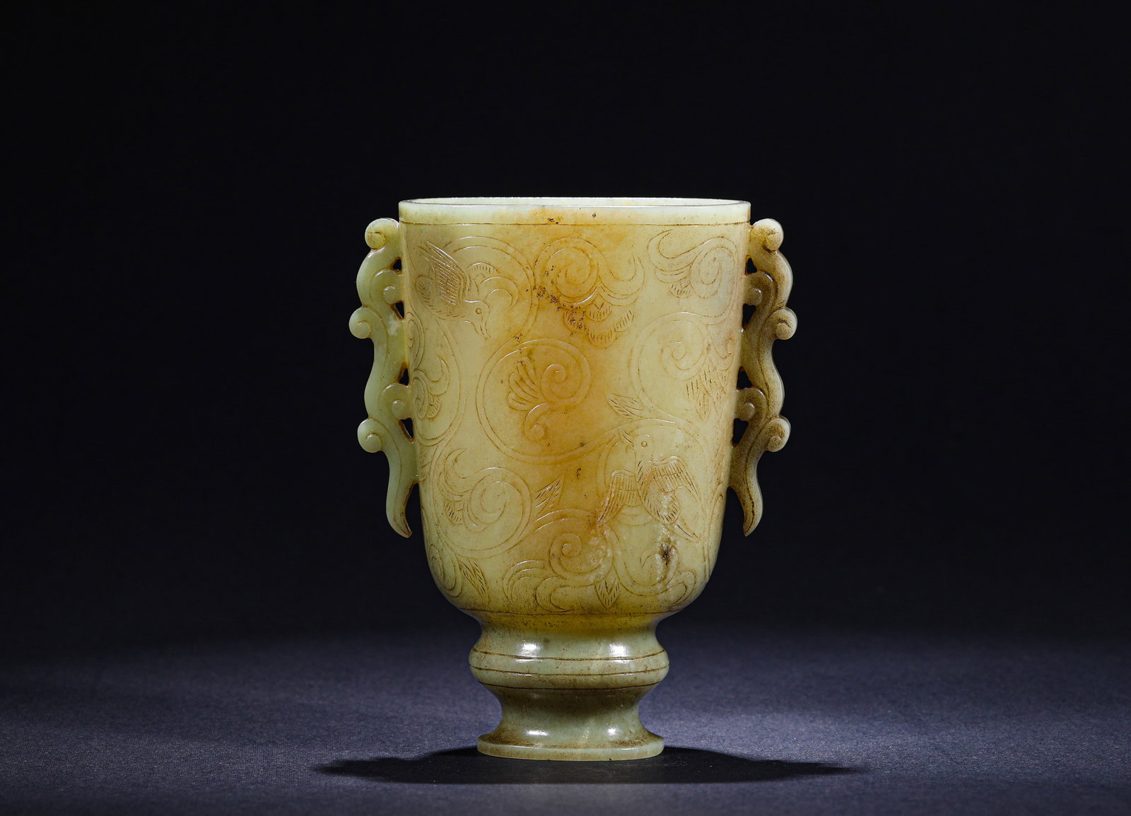 TANG DYNASTY GREEN AND YELLOW JADE SCROLL GRASS PATTERN DOUBLE-CHI EAR HIGH-FOOTED CUP: H:11.5cmW:9cmWeight:230g This is a tall-stemmed cup with double chi-dragon handles and scrolling grass patterns, carved from pale yellowish-green jade. The cylindrical body is flanked by openwork