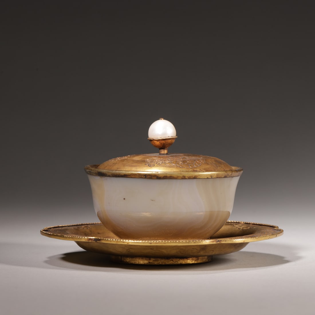An Agate Teacup with Gilt Copper Cover and Saucer, Qing Dynasty: D 12.5cm (4.92 in); H 8cm (3.15 in); Wt 290g (0.64 lb)A Qing Dynasty teacup set with an agate body, a gilt copper cover with a pearl finial, and a gilt copper saucer. The cover and saucer are engraved