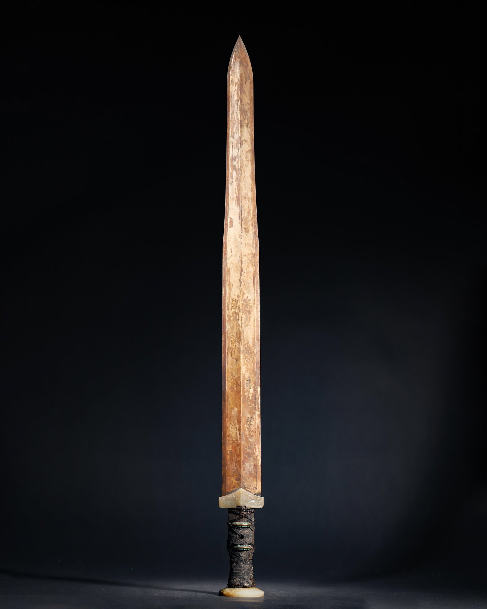 A Bronze ornament with Jade Guard and Pommel, Warring States Period: 1100.0 g (2.43 lb) | 70.0 x 5.5 cm (27.56 x 2.17 in)This Warring States bronze ornament with jade guard and pommel features a long, sharp blade with a distinct central ridge. The guard and pommel are 
