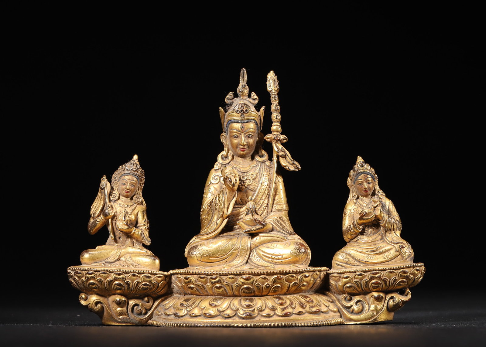 A Gilt-bronze Conjoined Statue of Padmasambhava and Two Attendants, Qing Dynasty: 667.0 g (1.47 lb) | 15.0 x 6.0 x 11.0 cm (5.91 x 2.36 x 4.33 in)This statue is crafted from fine bronze with rich, golden gilding. It features Padmasambhava in the center, holding a vajra and kartika,