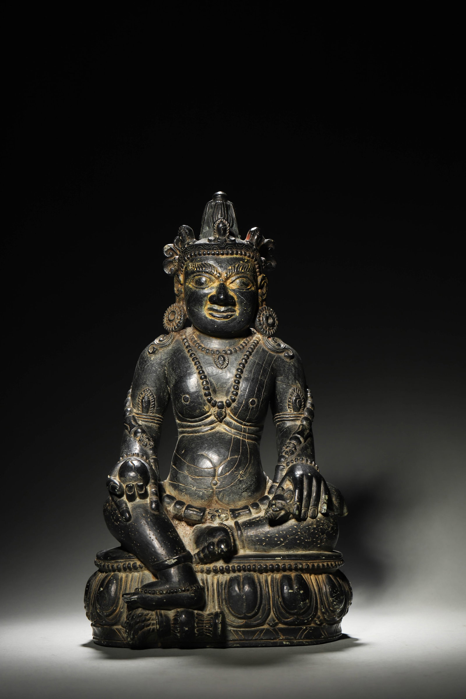 A Tibetan Black Stone Carved Statue of Jambhala, Ming Dynasty: L 18cm (7.09 in); H 34cm (13.39 in); Wt 6.85kg (15.1 lb)A Ming Dynasty Tibetan black stone carved Jambhala statue, with fierce yet compassionate expression, holding a wish-fulfilling jewel and a mongo