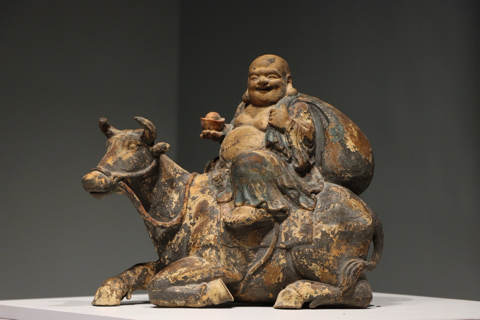 A Polychrome Wood Figure of Budai Riding an Ox - Qing Dynasty: Height 34cm (13.39 in); Width 37cm (14.57 in)This polychrome wood figure depicts Budai (the Laughing Buddha) seated on a reclining ox, dating to the Qing Dynasty. With a jovial expression, he holds an