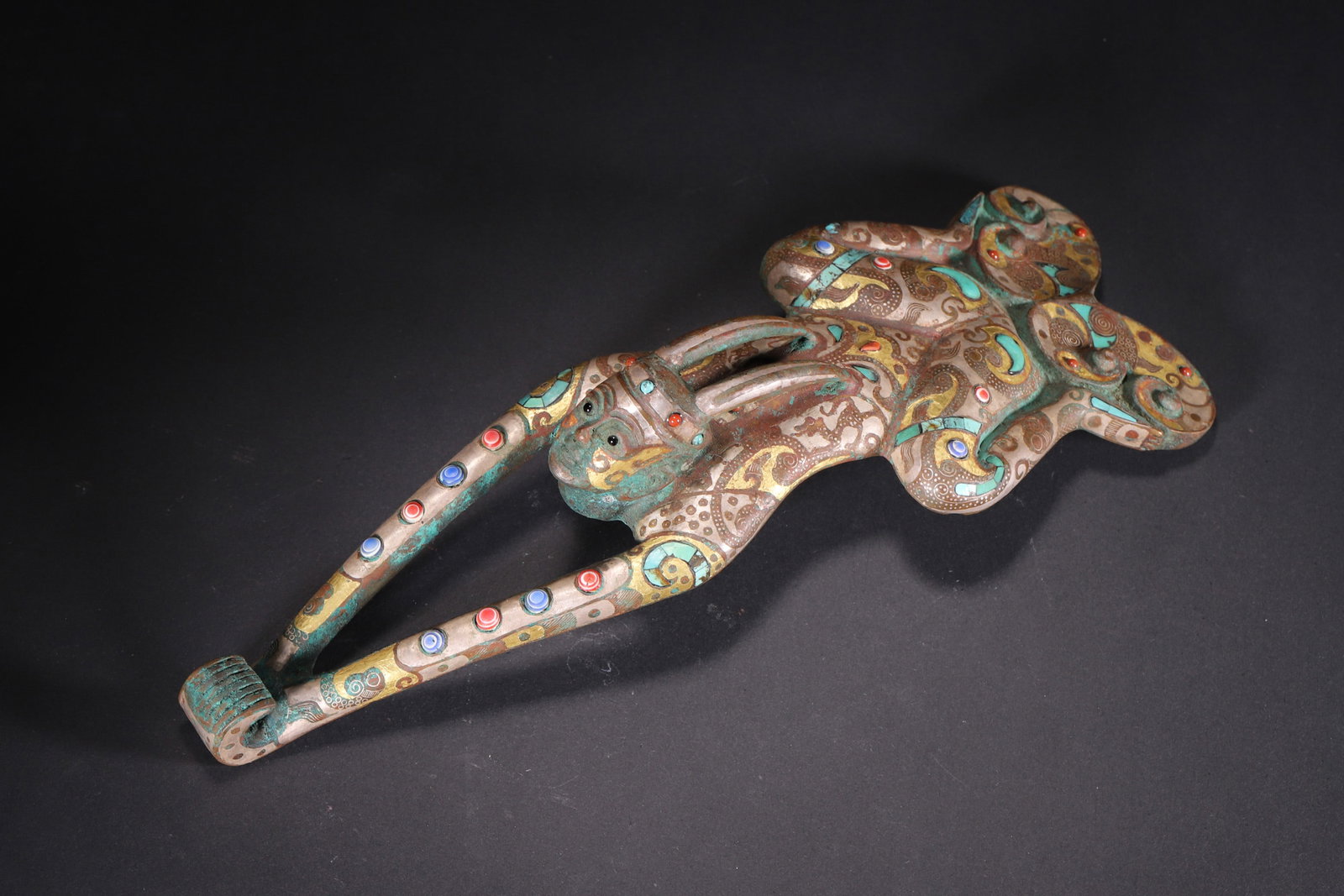 A Gold and Silver Inlaid Bronze Belt Hook with Multi-Gem Inlay and Deity Motif - Warring States: Length 27.0cm (10.63in), Width 9.0cm (3.54in), Weight 1102.0g (2.43lb)This Warring States bronze belt hook is shaped as a deity, inlaid with gold, silver, turquoise and multi-colored gems. The back fe