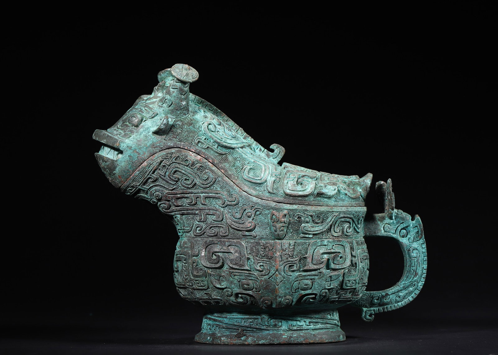 A Late Shang Dynasty Bronze Ritual Wine Vessel (Gong) with Taotie Pattern: 2427.0 g (5.35 lb) | 24.0 x 12.0 x 21.0 cm (9.45 x 4.72 x 8.27 in)This Late Shang Dynasty bronze ritual wine vessel (Gong) is robustly shaped with a beast-head lid. Its body is decorated with high-rel