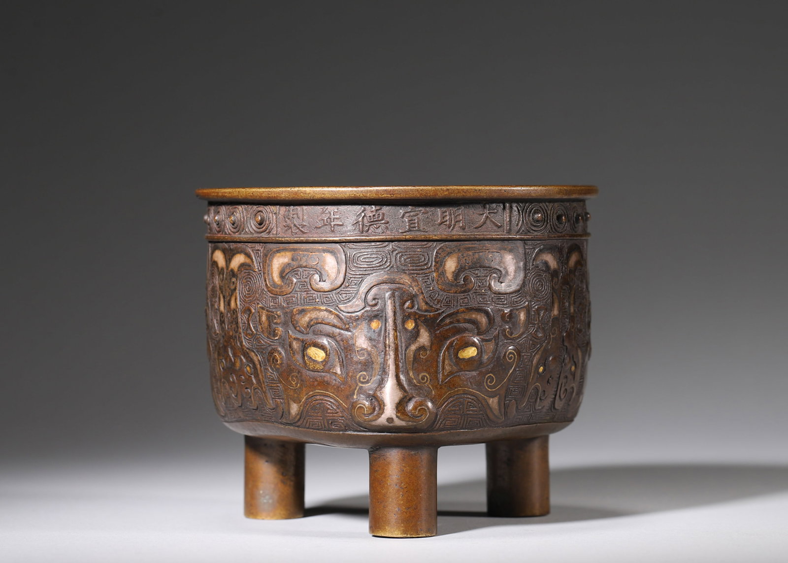 A Mixed Gold & Silver Inlaid Bronze Censer with Taotie Mask, Ming Dynasty: Dia 12cm (4.72 in); H 10cm (3.94 in); Wt 642g (1.42 lb)This well-cast Ming bronze censer bears six-character Xuande mark on rim. Its body is fully carved with archaic taotie patterns elaborately inlai