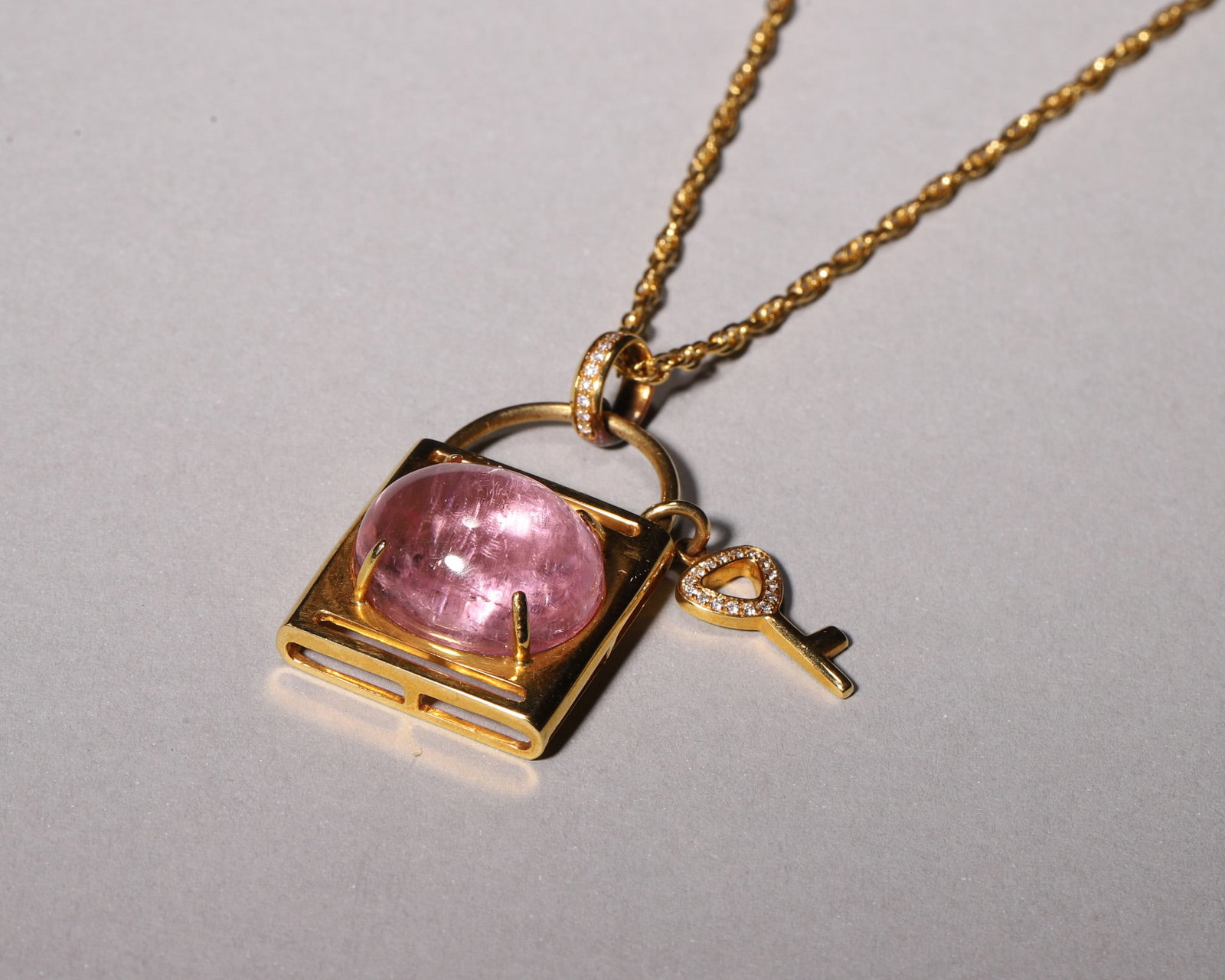 AN 18K GOLD LOCK-SHAPED TOURMALINE PENDANT NECKLACE WITH DIAMOND KEY CHARM: Wt 15g (0.033 lb);Worn vertical length L 25cm (9.84 in),Crafted in 18K gold, this lock pendant is set with cabochon pink tourmaline, paired with a diamond-set heart key charm. Matched with twisted rop