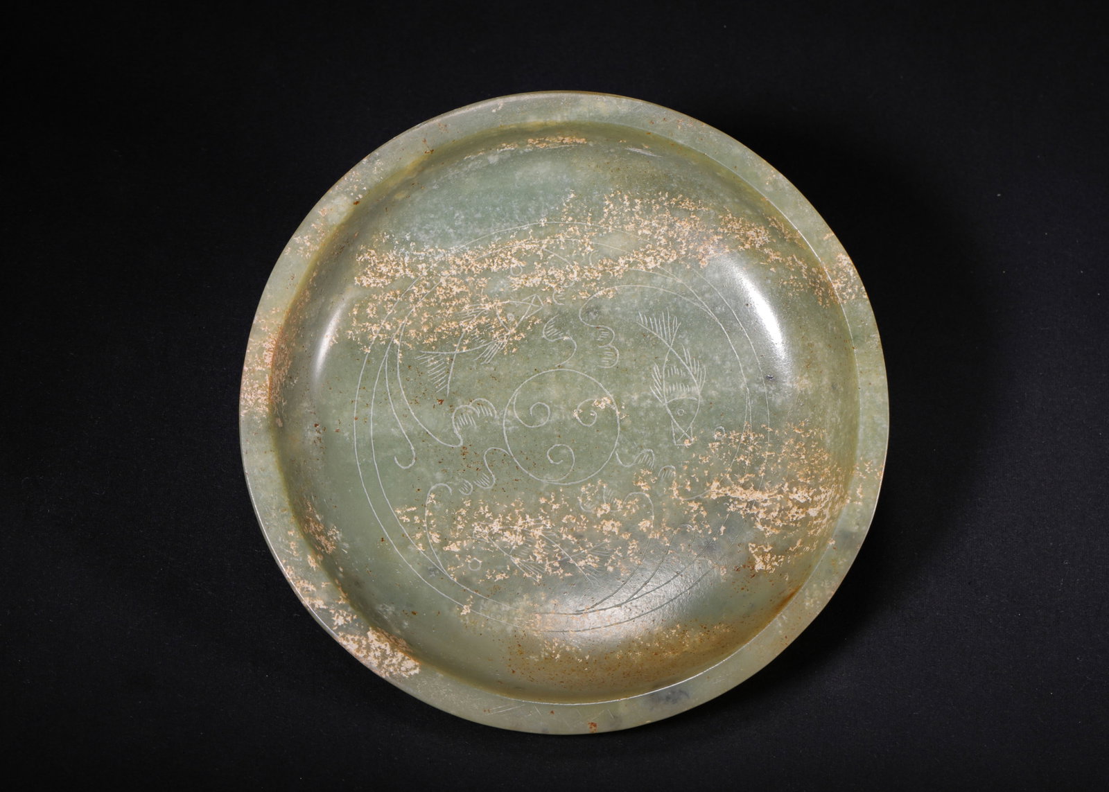 CELADON WHITE JADE DISH WITH FISH MOTIFS, HAN DYNASTY (202 BC-220 AD): H 3cm (1.18 in); D 14cm (5.51 in); Wt 215g (0.47 lb),Carved from celadon white jade covered with mottled earth encrustation. Shallow bowl with folded rim and ring foot. The interior is finely incised 