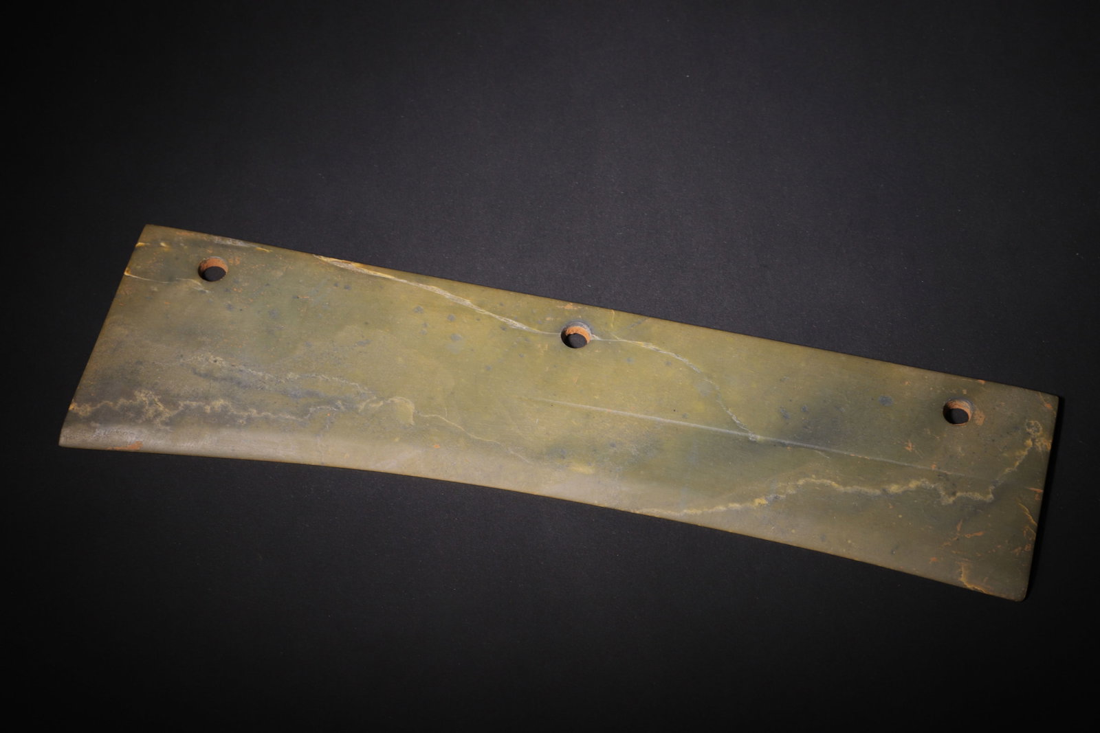 THREE-HOLE JADE KNIFE, SHANG DYNASTY (C.1600?C1046 BC): L 32cm (12.60 in); W 9cm (3.54 in); Wt 394g (0.87 lb),Carved from green jade naturally veined with dendritic inclusions and russet burial stains. Elongated flat curved knife form, three evenly spaced 