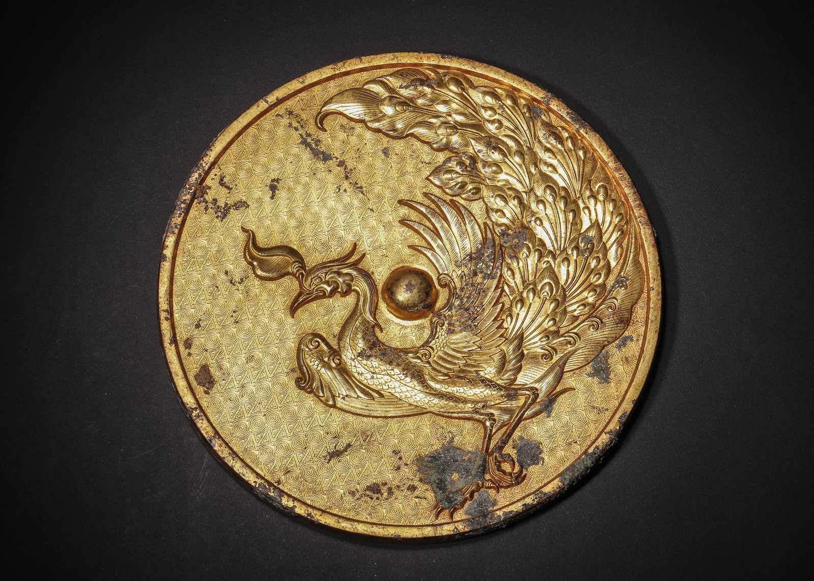 GILT BRONZE CIRCULAR MIRROR WITH PHOENIX FACING SUN MOTIF, TANG DYNASTY (618?C907 AD): D 20cm (7.87 in); Wt 1885g (4.16 lb),Cast bronze fully covered with gilding, the surface finely chased with repeating spiral ground pattern. A prominent high-relief phoenix facing a sun orb dominates 