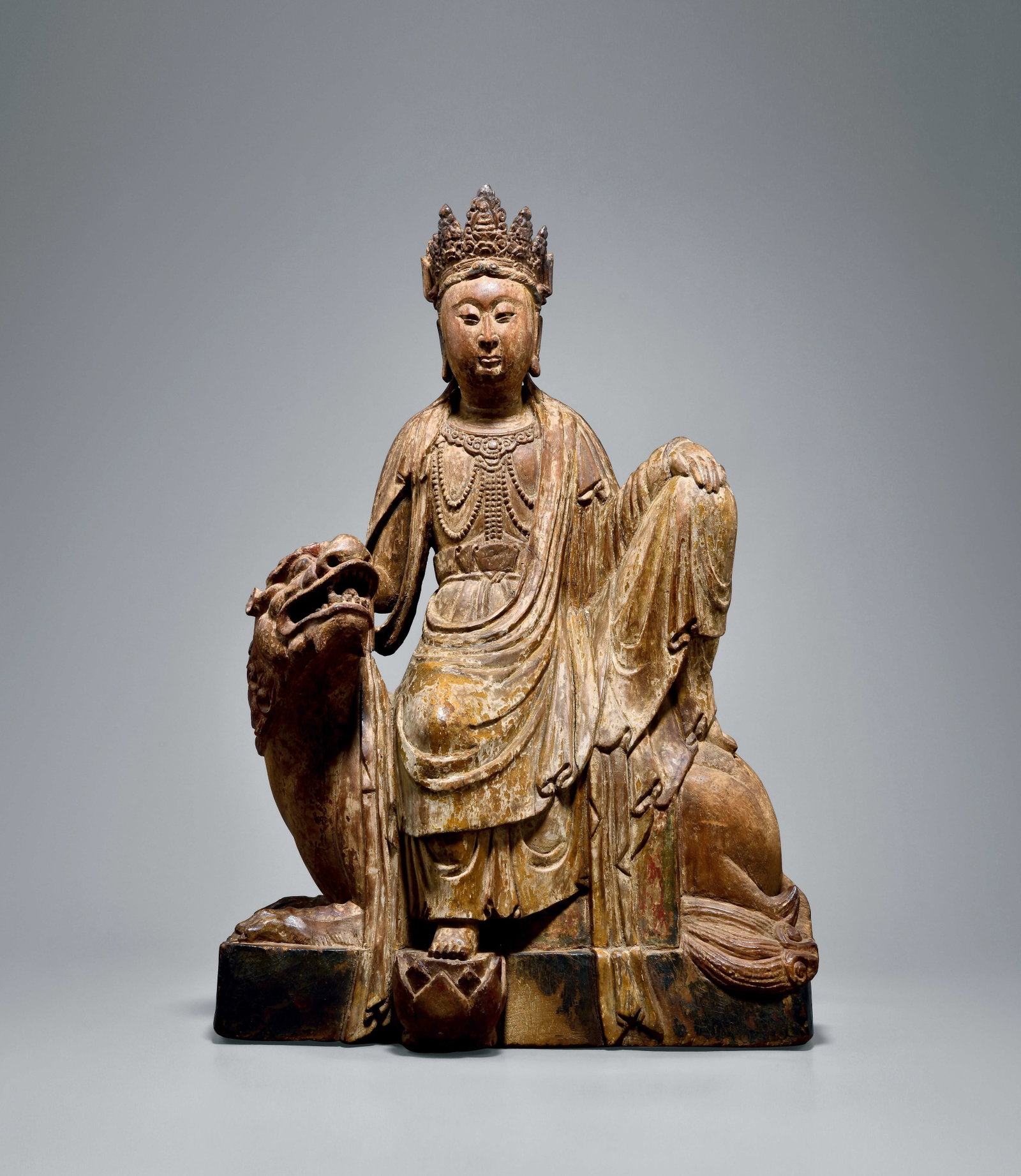 PAINTED WOOD SEATED SIMHANADA AVALOKITESHVARA, EARLY MING DYNASTY, 1368?C1450 AD: H 48cm (18.898 in),Simhanada Avalokiteshvara sits in relaxed posture atop a lion. The deity wears an ornate crown with serene, full facial features. Elaborate beaded necklaces adorn the chest; flowing