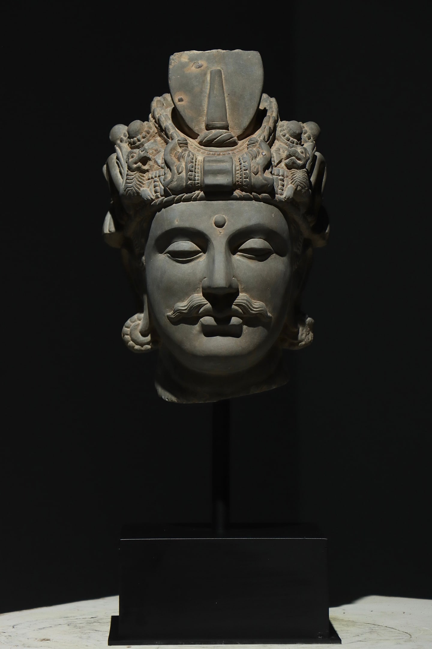 GREY SCHIST HEAD OF A MUSTACHIOED BODHISATTVA, GANDHARA, 2ND?C3RD CENTURY AD: Overall H 56cm (22.047 in); Net H 33cm (12.99 in),Carved wholly from grey schist in Hellenistic Gandharan manner. The face bears an urna on the forehead and defined moustache. The ornate crown is deco
