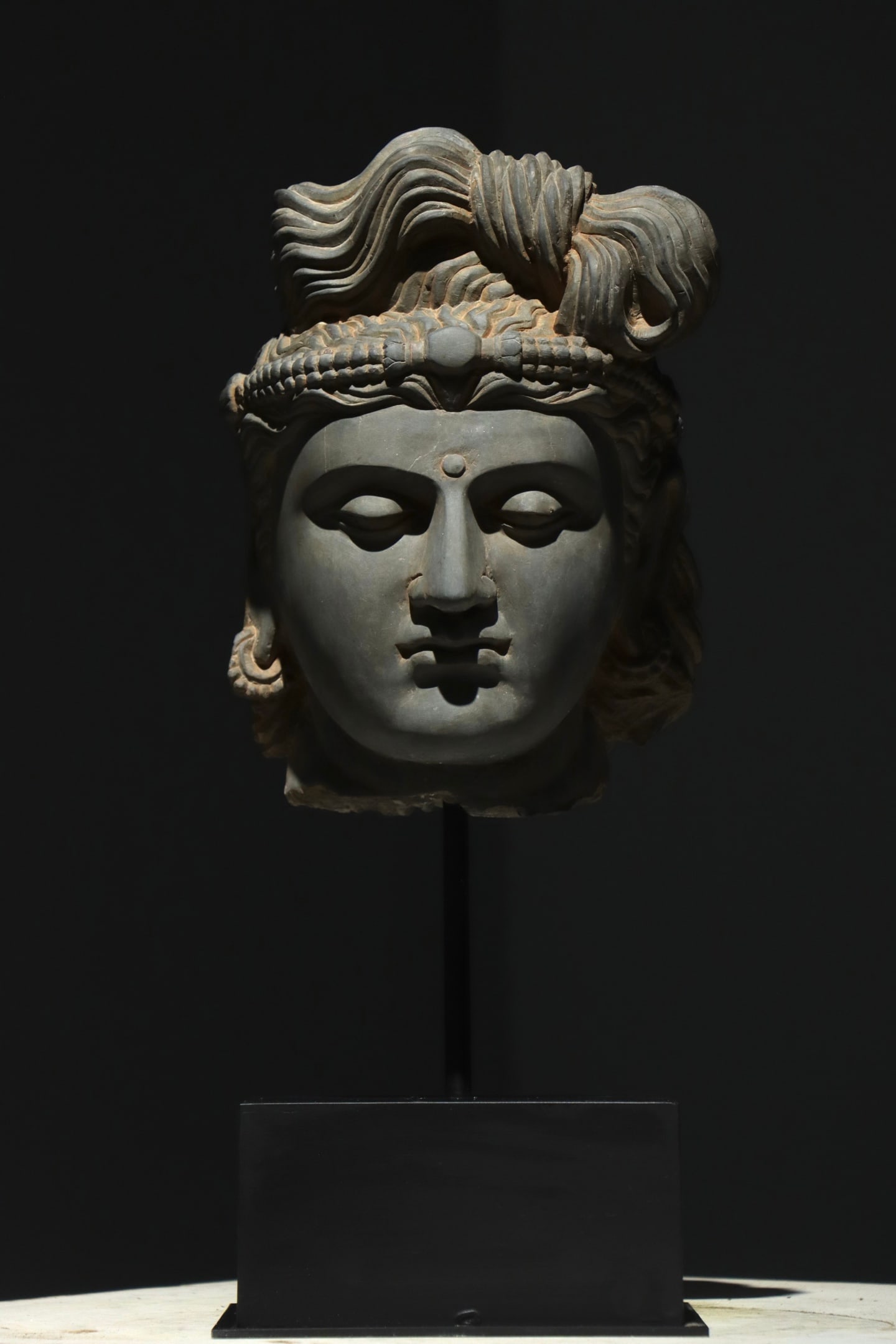 GREY SCHIST BODHISATTVA HEAD, GANDHARA, 2ND?C3RD CENTURY: Overall H 50cm (19.69 in); Net H 30cm (11.81 in),Grey schist carved bodhisattva head with broken neck. Hair coiled into a high topknot secured by a beaded diadem, finely rendered wavy locks. Face exec