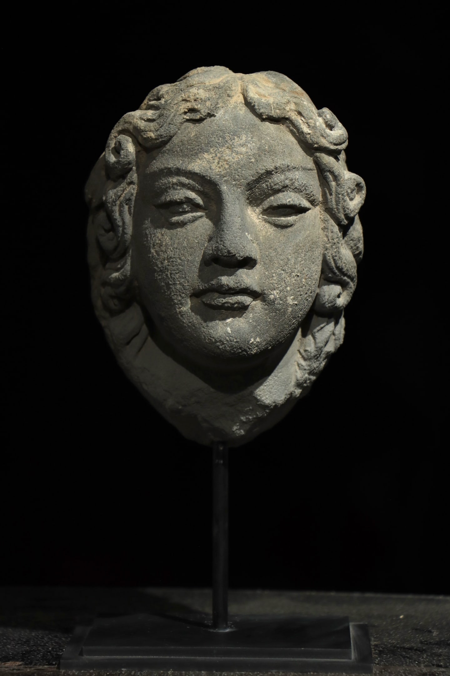 Grey Schist Head Of Devata, Gandhara (2Nd 3Rd Century Ad): Overall H 28cm (11.024 in); Net H 19cm (7.48 in)Carved from grey schist, this devata head features tightly coiled spiral curls. Its facial characteristics combine Hellenistic realism with Eastern aest