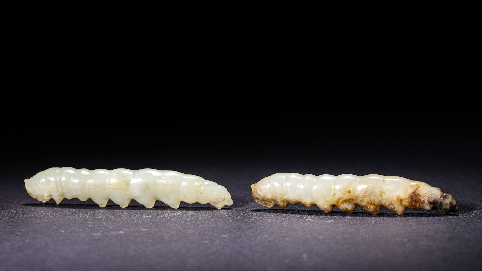 A Pair Of Hetian Jade Silkworm Larvae, Han Dynasty: Wt 34g (0.075 lb); L 7.2cm (2.83 in); W 1.4cm (0.55 in)A pair of circular carved Hetian celadon jade silkworm larvae. One shows brown black natural patina. Segmented bodies with carved abdominal feet,
