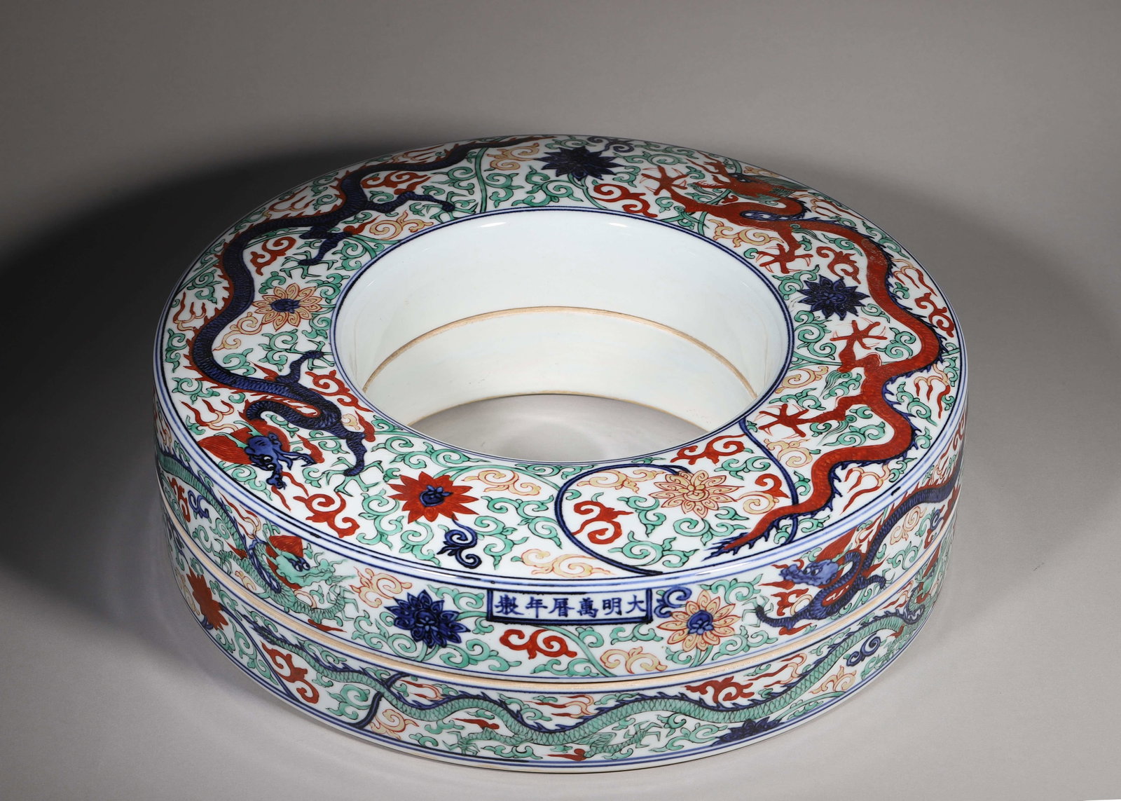 Wucai Circular Ringshaped Covered Box With Dragon Scrolls, Wanli Ming (15731620 Ce): W 40cm (15.75 in); H 12cm (4.72 in)Circular ringshaped covered box with fitted joint. Fully decorated in wucai with dragons winding through scrolling lotus blossoms. Underglaze blue Wanli reign mark o