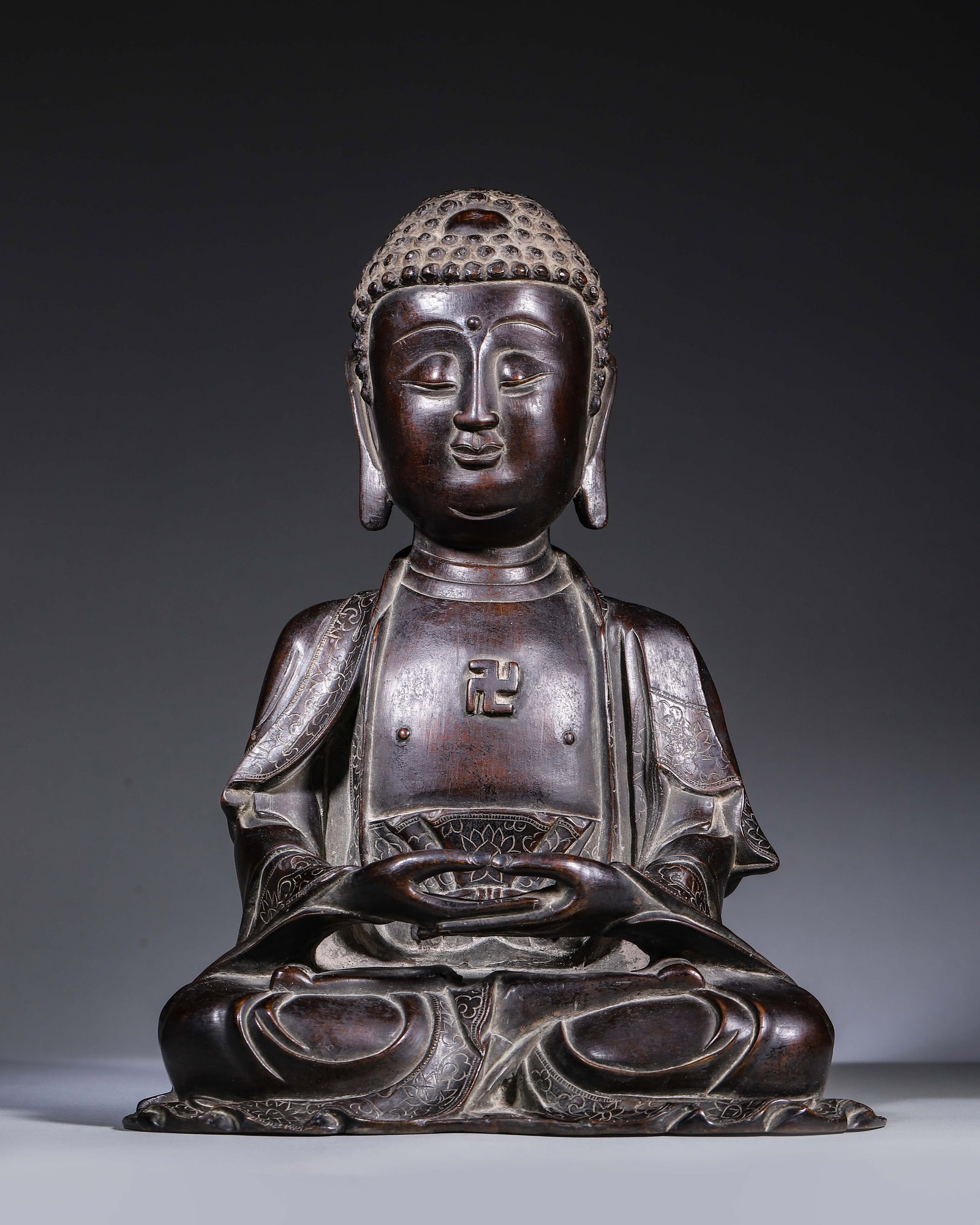 Ming Dynasty, Bronze Seated Sakyamuni: This bronze Sakyamuni sits in full lotus posture with dhyana mudra. It features regular spiral hair, a serene countenance, a swastika symbol on the chest, and lotus patterns engraved on the robe edge.