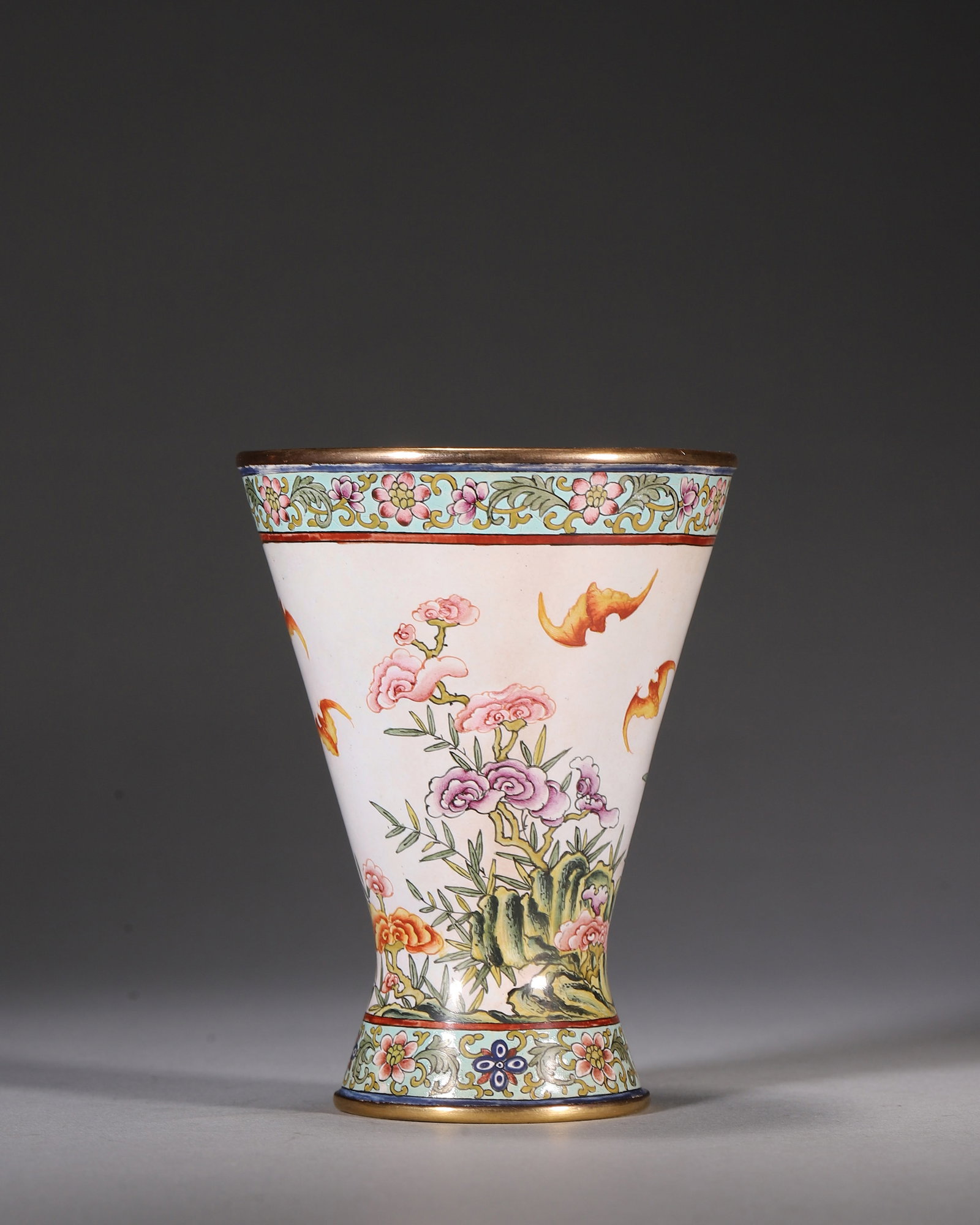 Bronze-Bodied Painted Enamel Flared Cup With Bats, Flowers And Rocks, Qianlong Period, Qing Dynasty: Gilt-bronze body. White ground decorated with lingzhi, flowers, rocks and flying bats symbolizing blessing and longevity. Floral scrolls adorn rim and base, gilt rim and foot. Base bears blue enamel "