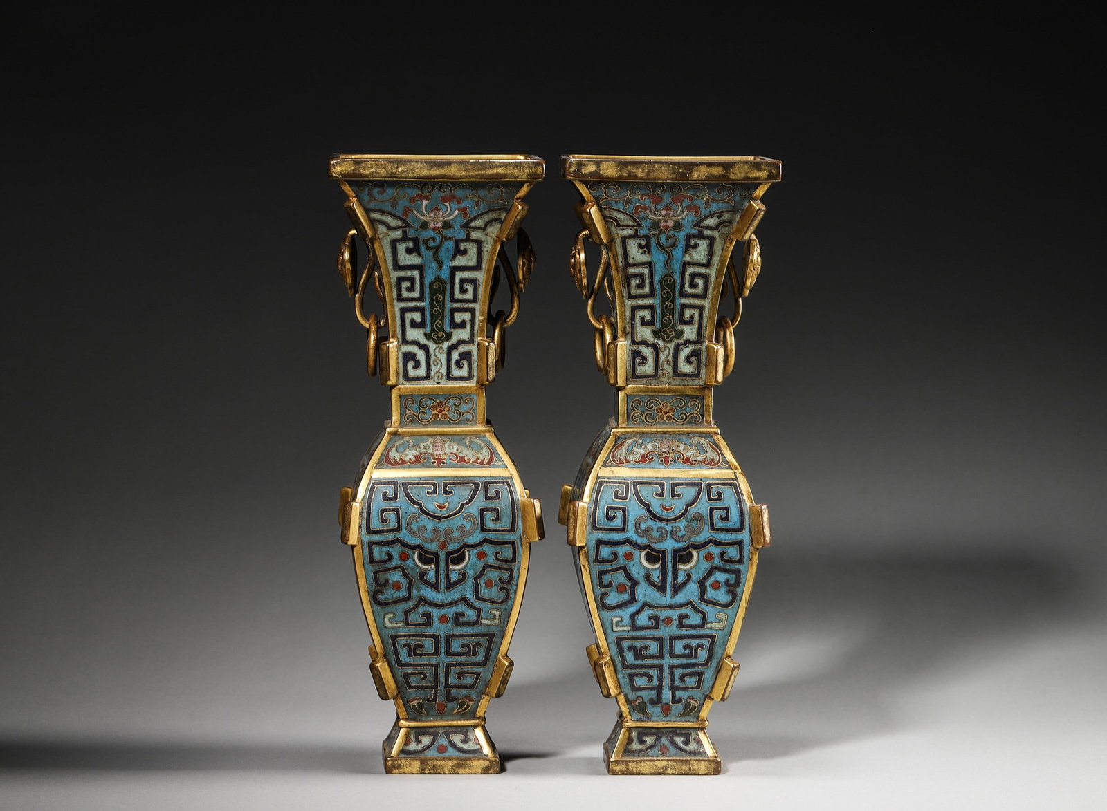 Pair Of Qianlong Period Cloisonne Enamel Archaistic Square Vases: Pair of cloisonne vases modelled after archaic bronze form, beast-head loose ring handles, mask and bat motifs on turquoise ground, gilt flanges, incised Qianlong mark at base, imperial pair display p