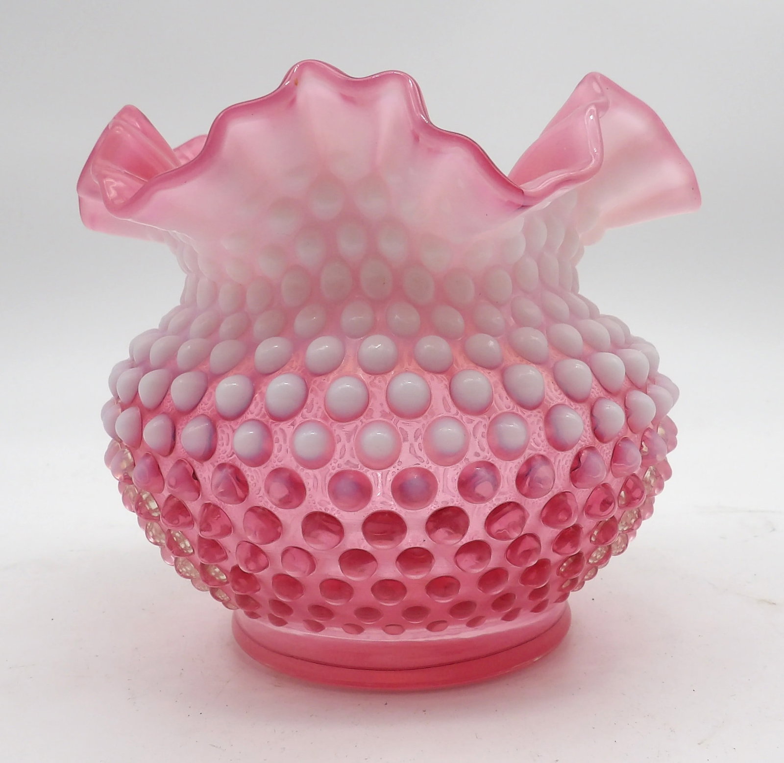 FENTON CRANBERRY OPALESCENT HOBNAIL RUFFLED RIM VASE: This vase is characterized by its cranberry color, hobnail pattern, and ruffled rim. It is a great piece for collectors of antique, vintage, and art-style glass. (5” X 6”)