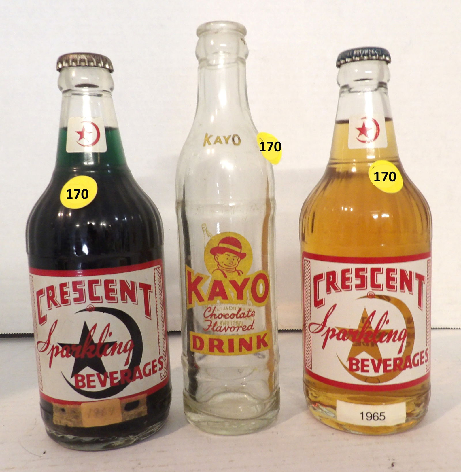 Crescent Grape, Crescent Cream Soda, Kayo Glass Bottles (3 pcs): This group of vintage glass soft drink bottles includes a 1964 Crescent Grape (sealed w/ original contents), a 1965 Crescent Cream Soda (sealed w/ original contents), and a 40's / 50's Kayo Chocolate