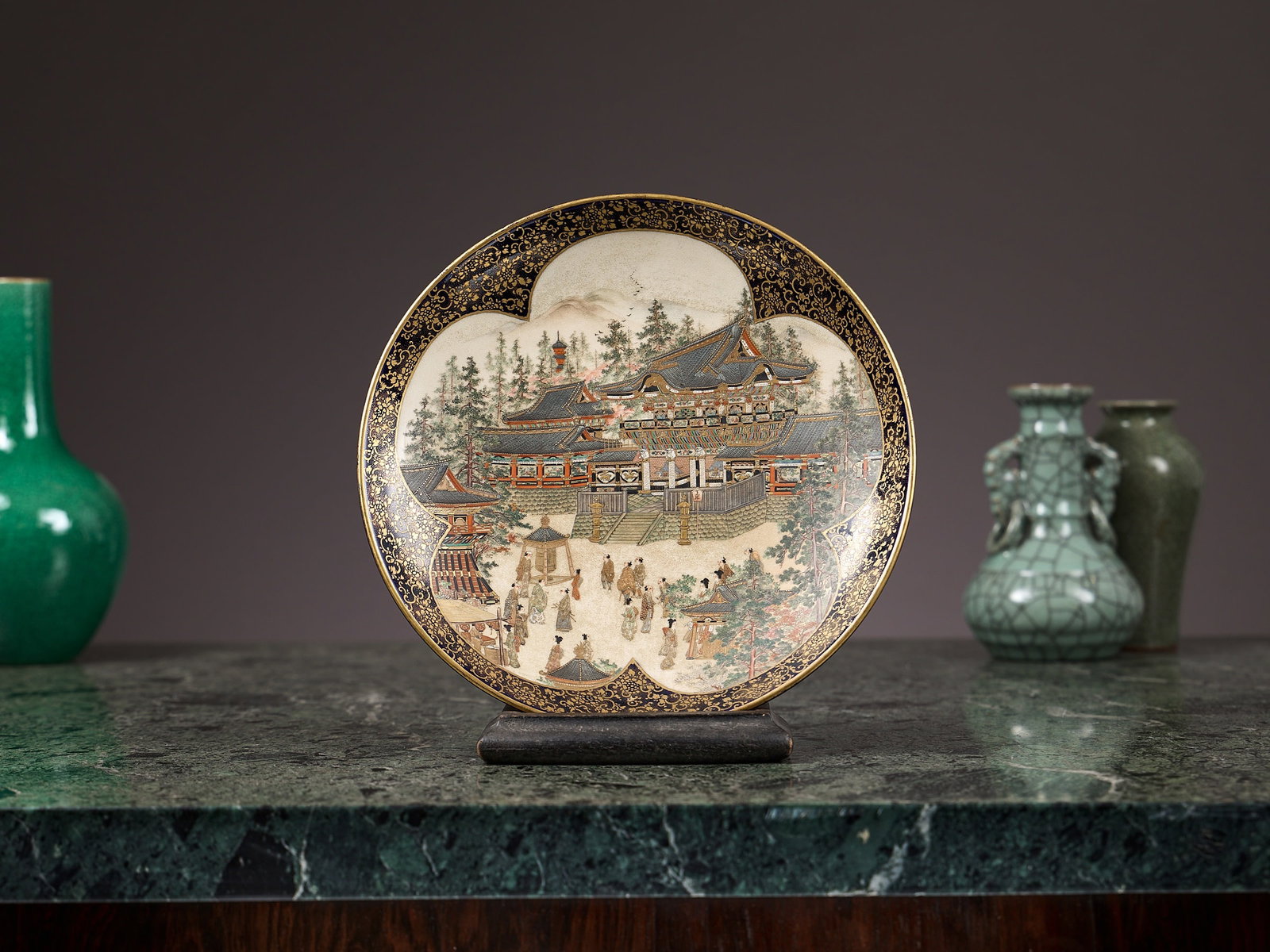 A JAPANESE MEIJI PERIOD SATSUMA WARE PLATE: A JAPANESE MEIJI PERIOD SATSUMA WARE PLATE by Kinkozan, the ceramic dish of circular form, painted in a prunus shaped panel with figures and buildings, within a border of gilt flowers on dark blue gro