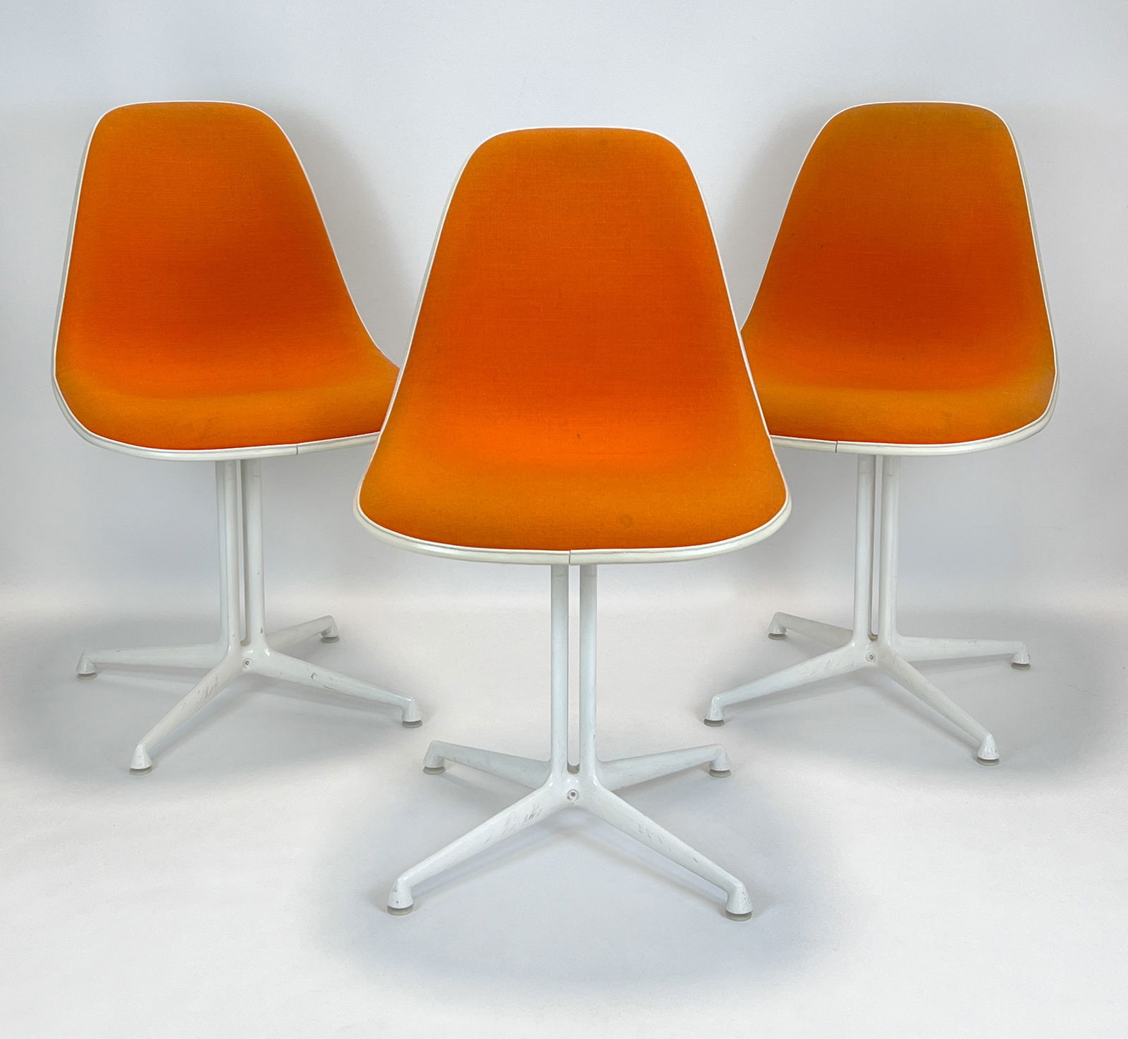 Charles & Ray EAMES (20th–21st centuries). Three ‘La Fonda’ chairs. Hermann: Approx. 80 cm x 47 cm x 40 cm. Seat height approx. 45 cm. Set of 3 ‘Side Chair / La Fonda’ chairs. Fibreglass seat shells with orange hopsack/fabric cover. Cast aluminium frame with white