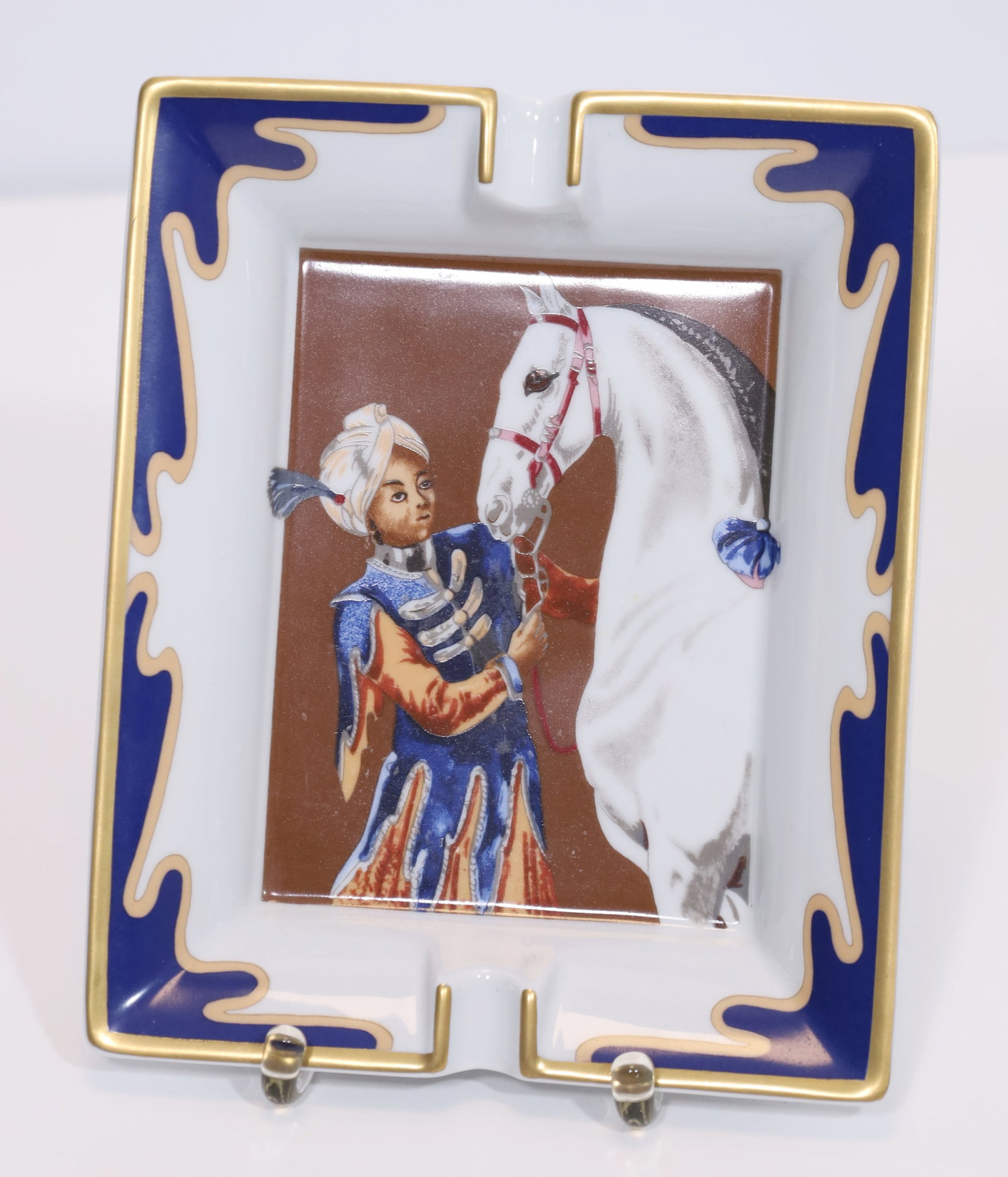 HERMÈS. Ashtray. ‘Sultan with white horse’.: Approx. 4 cm x 19.5 cm x 16.5 cm. Porcelain. Rectangular, recessed shape. 2 cigarette holders on the sides. Polychrome depiction of a sultan with a white horse in the centre. Gold decoration. Marked &