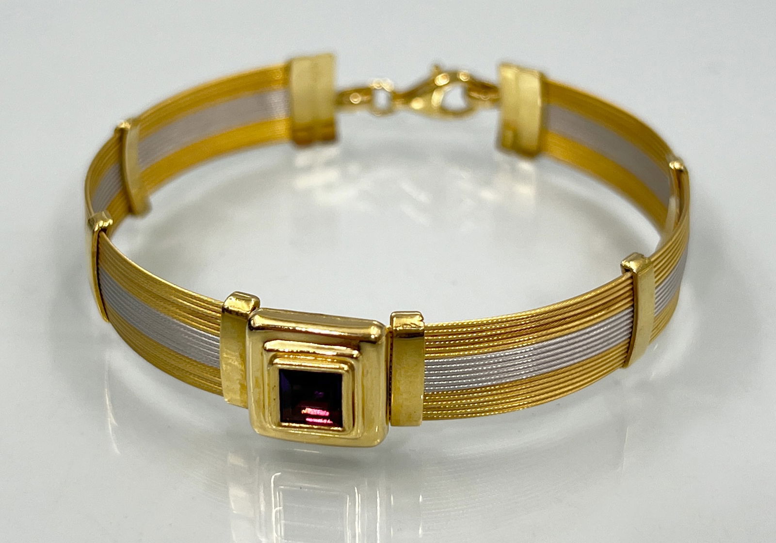Two-tone bracelet. 750 yellow gold and white gold with a purple gemstone.: Approx. 15.4 grams total weight. Approx. 17 cm long when open. Band finely crafted with thread detailing. One square-cut coloured stone (probably amethyst) measuring approximately 5 mm x 5 mm in the c