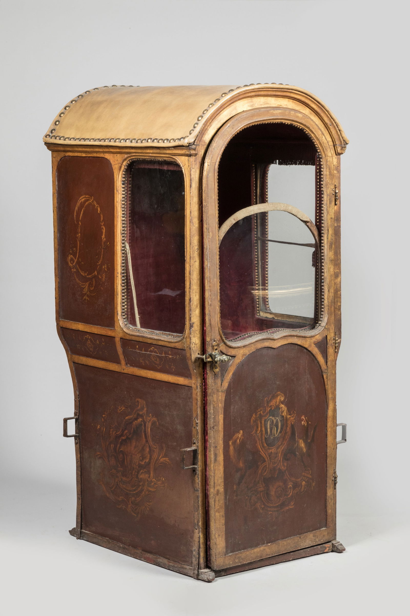 CHAISE A PORTEUR en bois moulure a decor peint de blason, chiffre D et coquille sur fond marron.: CHAISE A PORTEUR en bois moulure a decor peint de blason, chiffre D et coquille sur fond marron. Elle ouvre par une porte et presente trois fenêtres en partie haute; le toit en cuir bombe a decor clo