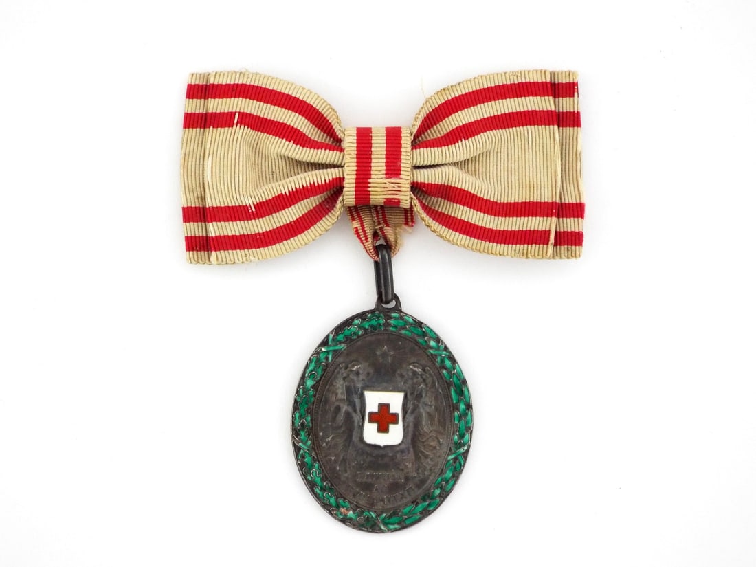 Austro-Hungarian WWI RED CROSS MERIT MEDAL: Silver toned metal body, obverse base reads PATRIAE AC HUMANITATI - For Fatherland and Humanity. Reverse displays relief dates 1864 1914 within a beaded border. The green enameled oak and laurel wreat