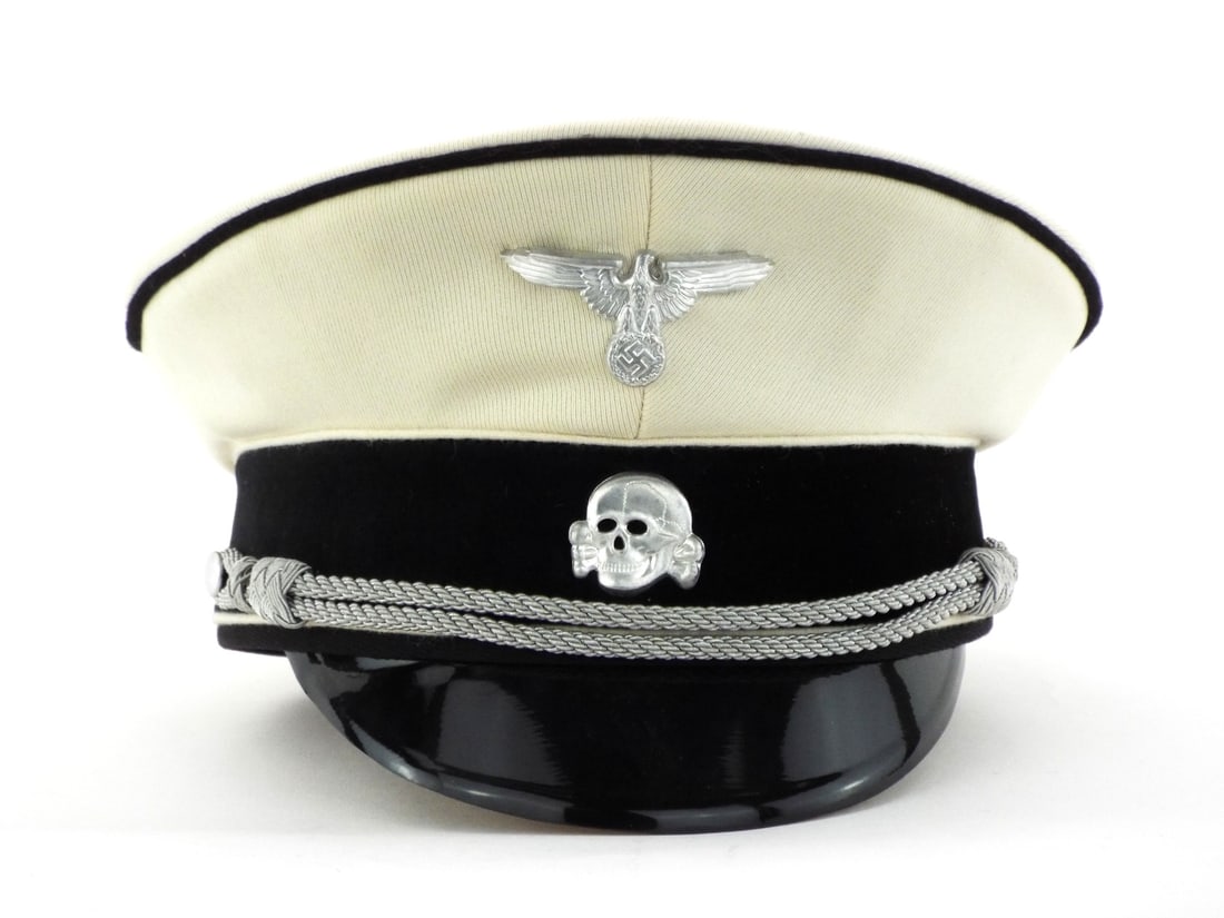 Very Good Quality German Made POST WWII SS Officer Visor Cap: White tricot wool body exhibits tight weave with black velvet band and black piping. Aluminum eagle and death head insignia show minimal oxidation. Interior features tan leather sweatband with perfora