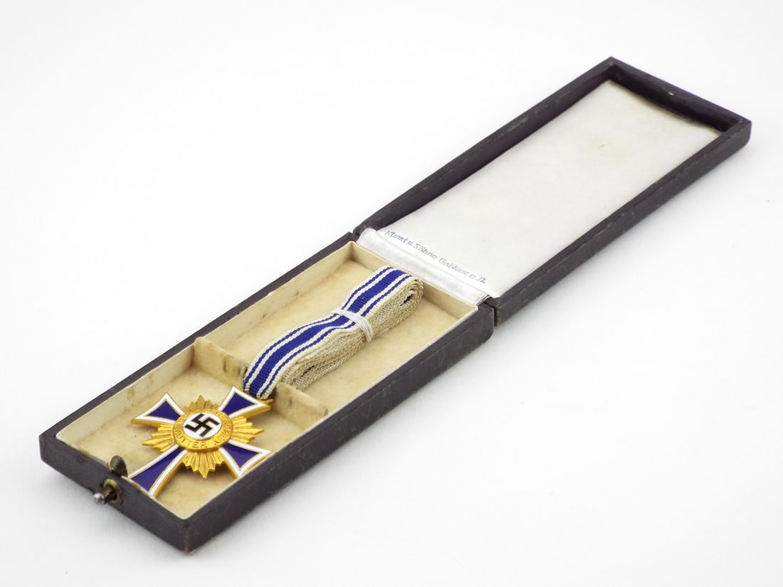 German WWII Encased Mother Cross Medal By KLAMT & SOHNE with Enamel & Gold Finish: Exhibits a bright gilded cross body with very minor handling wear to the blue and white enamel, flanked by intact sunburst rays. Suspended from its intact blue and white striped ribbon showing light a