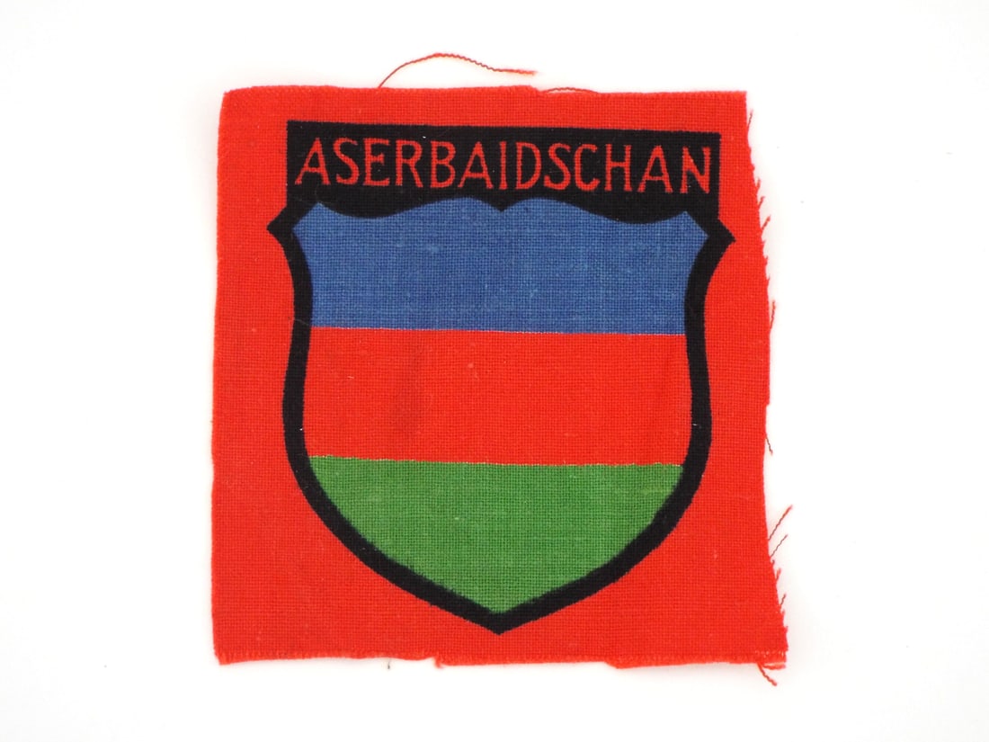 German WWII AZERBAIJANI VOLUNTEER LEGION Printed Cloth Sleeve Patch: Screen printed cotton construction patch features a tricolor blue, red and green shield beneath black lettering translating to Azerbaijan. Unissued condition displays light fraying along raw unhemmed 