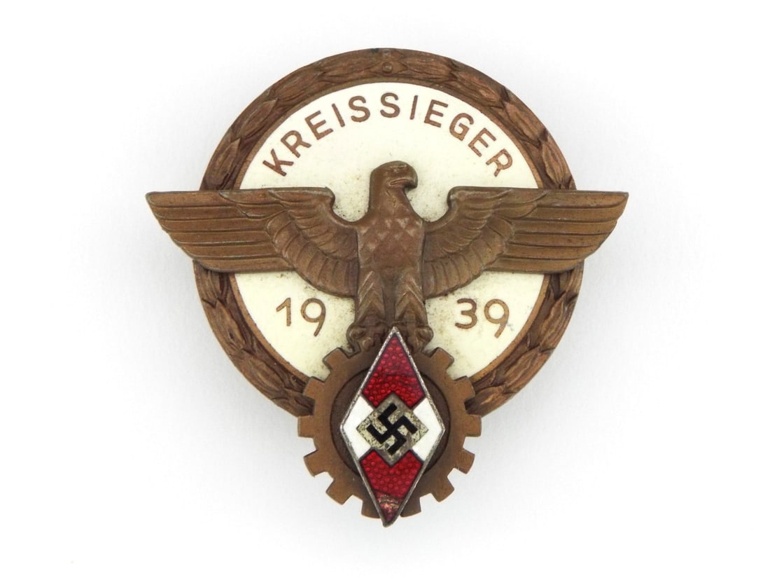 German WWII District Winner Badge MAKER FERDINAND WAGNER Enamel Field: Multi piece bronze construction award badge exhibits fine detail to central eagle, cogwheel and diamond emblem with intact red and white enamelwork showing light surface wear and minor scratching. Rev