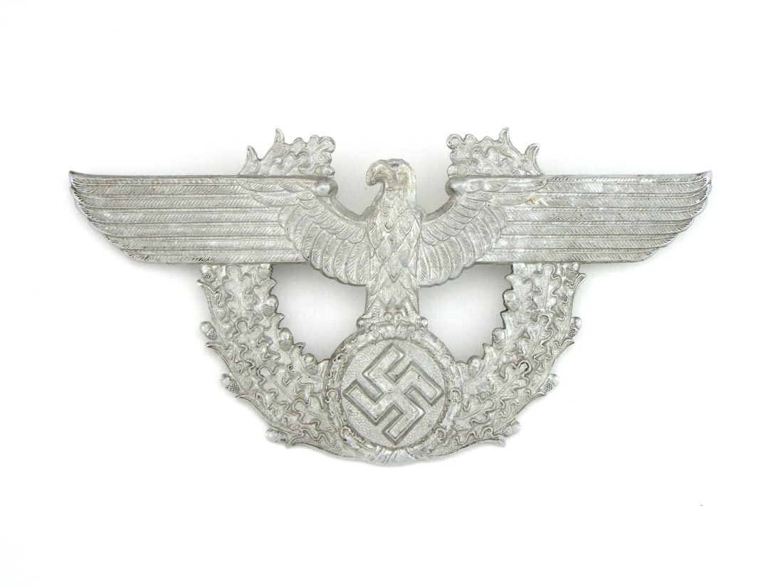 German WWII Police Shako Badge In Silvered Alloy: Stamped body features an eagle with outstretched wings clutching a oak leaf wreath enclosing a central mobile motif and soft wear to high points. Reverse retains crisp structural detail throughout wit