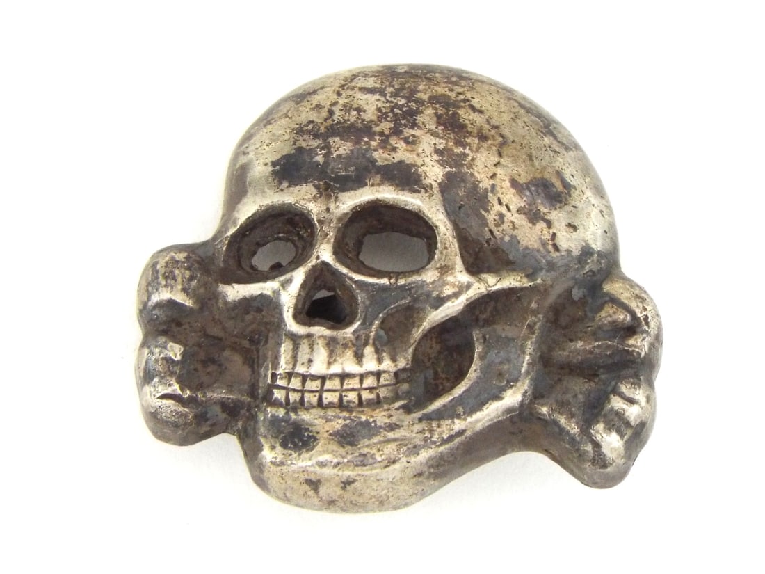 German WWII SS Visor Cap SKULL Insignia with RZM Number: Metal skull emblem displays silvered surface tones with prominent dark toning throughout the face contours and recesses. Reverse hollow cavity exhibits cast stippling alongside raised RZM symbol, SS 3