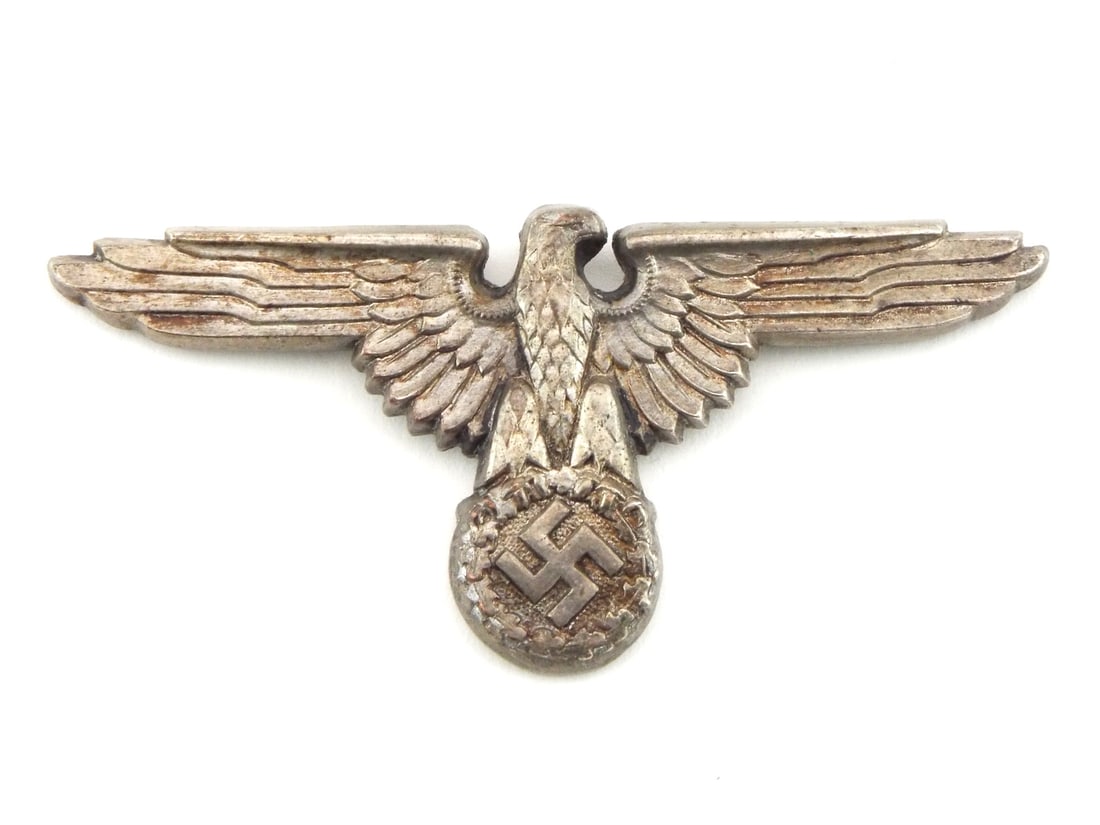 German WWII SS Cap Eagle By ASSMANN LUDENSCHEID RZM Number: Insignia body presents stamped die cast metal construction featuring open winged eagle motif above oak leaf wreath and mobile sunwheel symbol. Reverse displays hollow backed casting with integrated mo