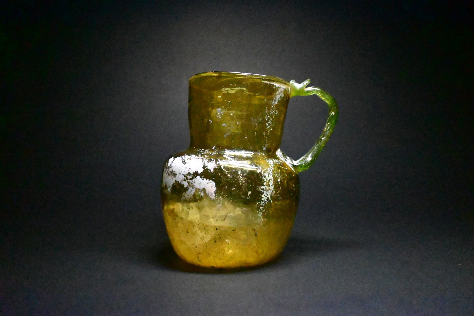 An islamic abbasid glass pitcher: Ancient Islamic Abbasid yellowish-green glass pitcher or jug, dating to the 8th–10th century CE. The vessel is blown in translucent olive-green glass with a globular body, tapering cylindrical neck,