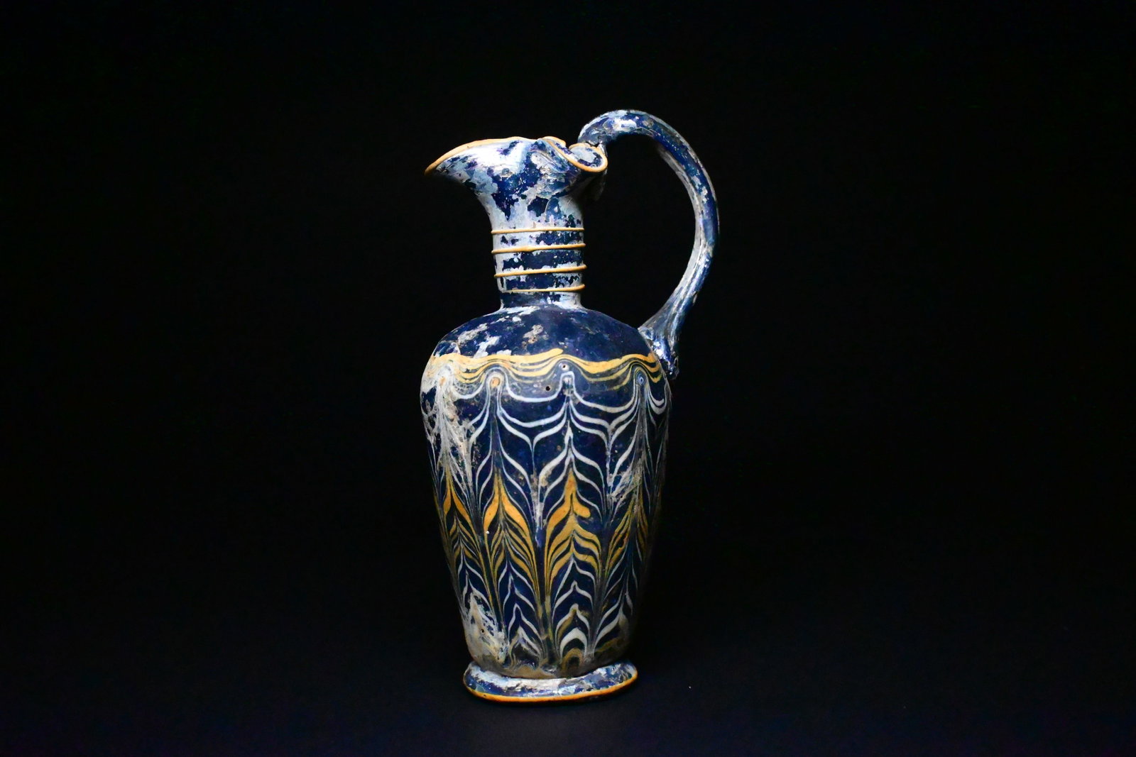 A Hellenistic CORE-FORMED GLASS OENOCHOE: circa mid 4th-early 3rd century b.c. hellenisticDark blue in color, the cylindrical body with broad, rounded shoulders, on a circular pad base, the cylindrical neck with trefoil rim and a vertical str