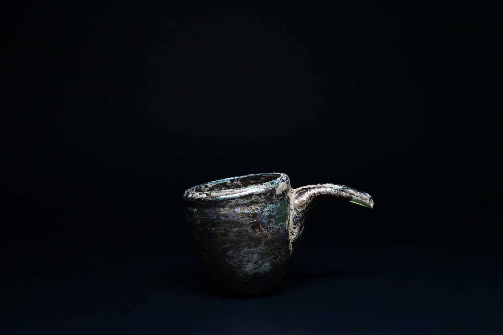 A roman glass feeder: A rare blown Roman glass feeder vessel of elegant miniature form, featuring a rounded cup-shaped body with a long curved tubular spout designed for controlled pouring or feeding. Circa 1st-4th Century