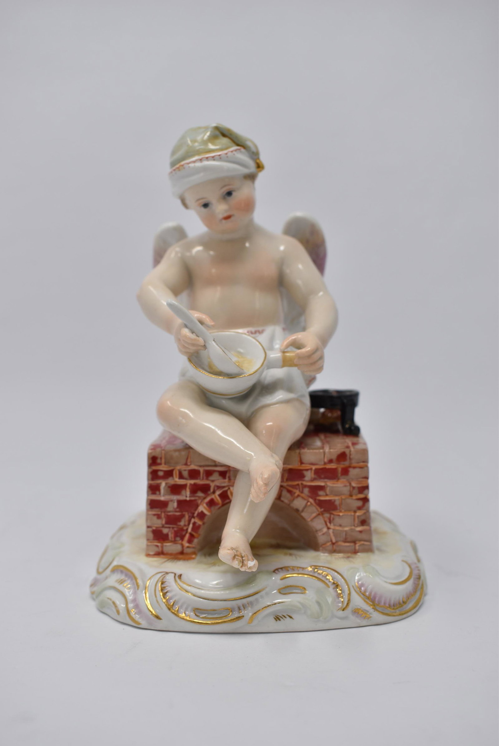 Meissen porcelain cherub eating porridge: A German porcelain figurine of a cherub seating on a brick stove eating porridge. Underglaze blue marks for Meissen. Approximate measurements: 6"H x 4"W x 3.5"D