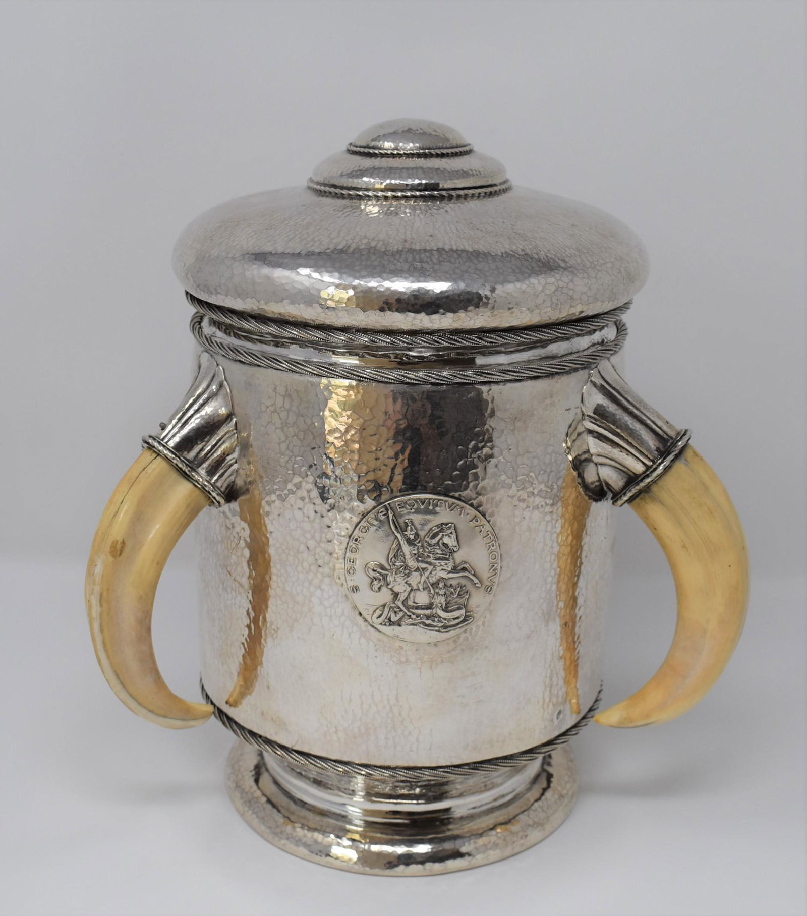 Antique German sterling silver trophy cup and cover: An antique German .935 sterling silver presentation trophy cup and cover with triple boar's tusk handles, martele finish and gilt interior. The applied central medallion depicting St george and inscri