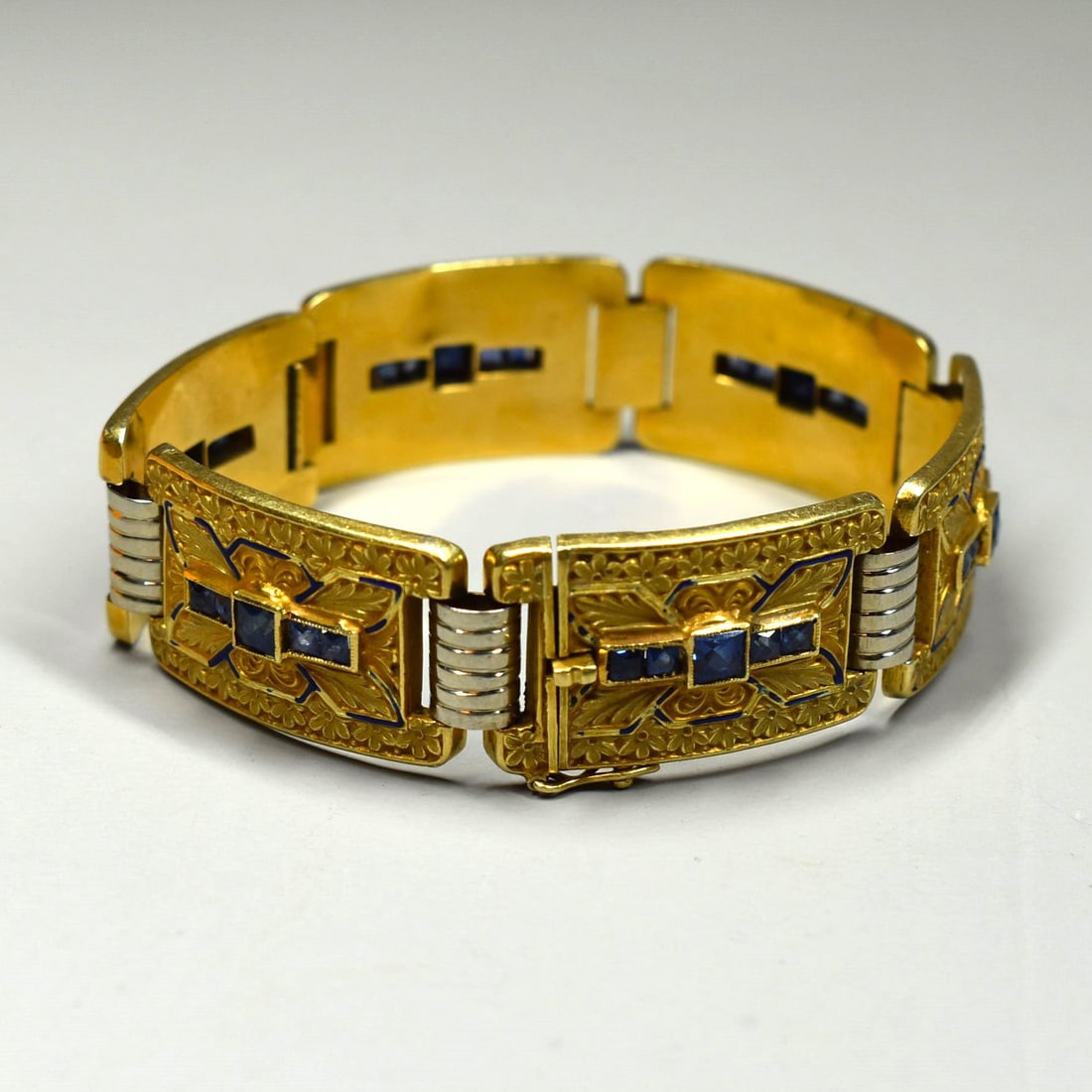 A French Art Deco 18K yellow gold bracelet with enamel and sapphire accents: A French Art Deco period (1930's/ 1940's) 18K yellow gold bracelet with blue enamel and sapphire accents. The large rectangular links with embossed decoration featuring square-cut sapphires. Marked '1