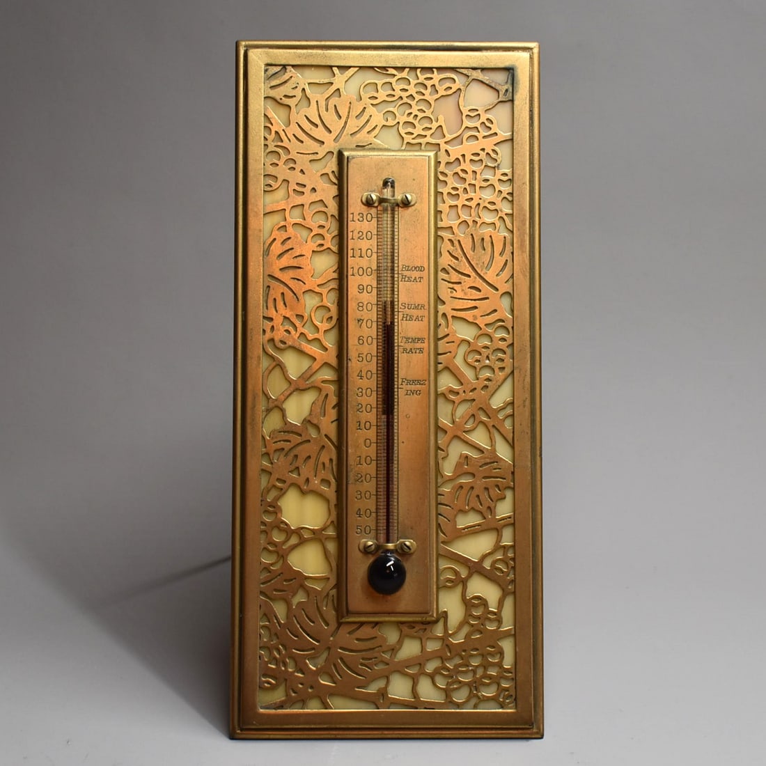 TIFFANY STUDIOS gilt bronze 'Grapevine' thermometer #1013: GILT BRONZE TIFFANY STUDIOS "GRAPEVINE" THERMOMETER: Tiffany Studios, #1013, Filigree Grapevine motif Gilt Bronze frame with a beaded border. Caramel slag glass insert. Impressed stamp at lower revers